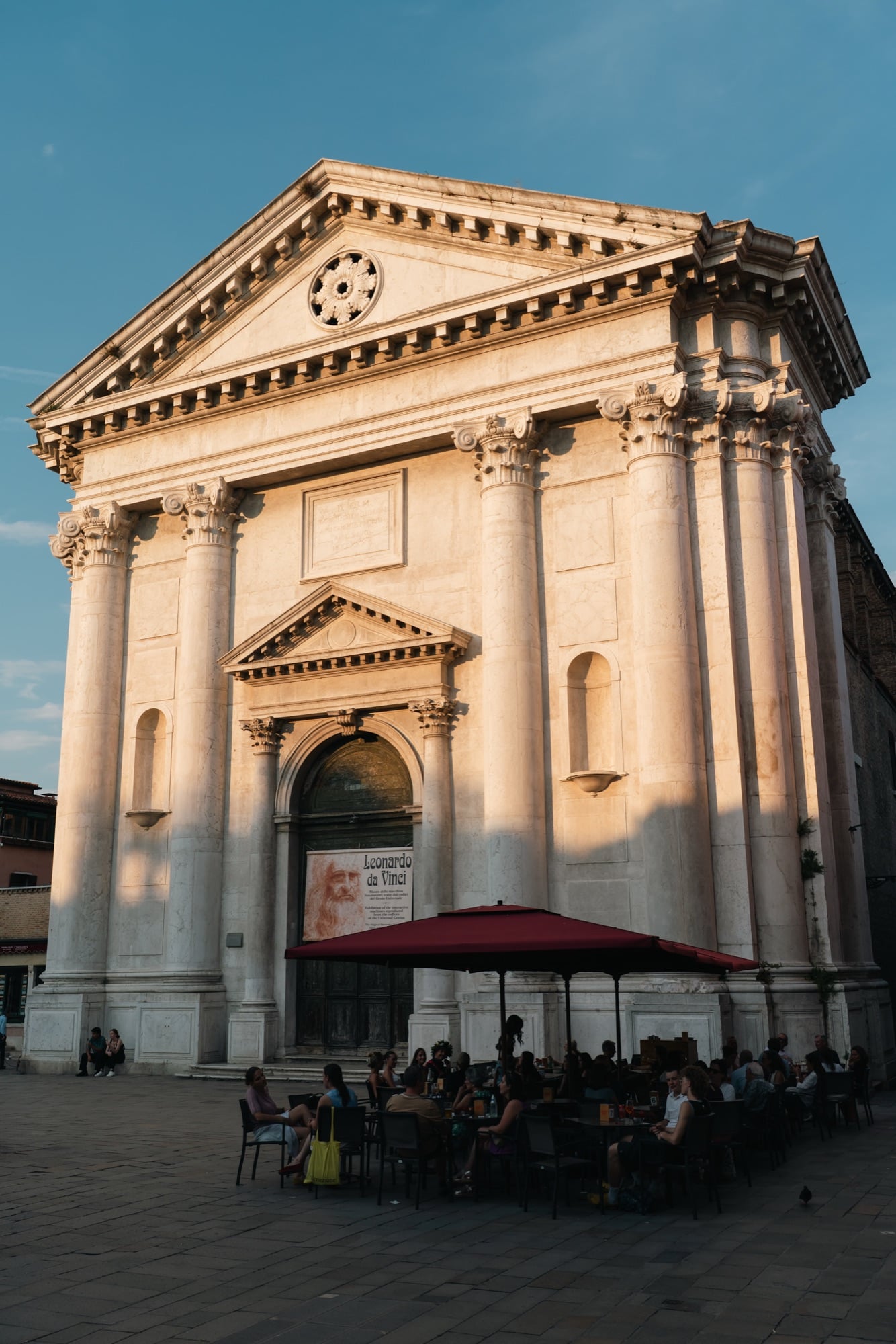 The Palladian columned front of the San Barnaba church glowing gold, a Leonardo da Vinci exhibition banner over the door and diners at café tables below