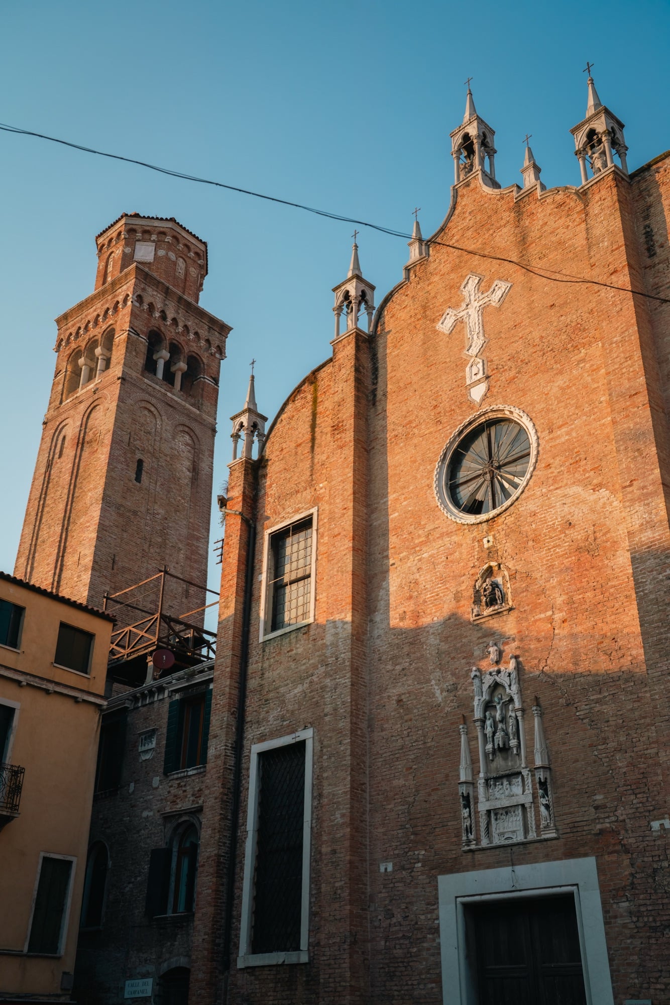 The tall brick campanile and Gothic apse of the Frari basilica lit gold by low sun, a white marble crucifix on the brickwork
