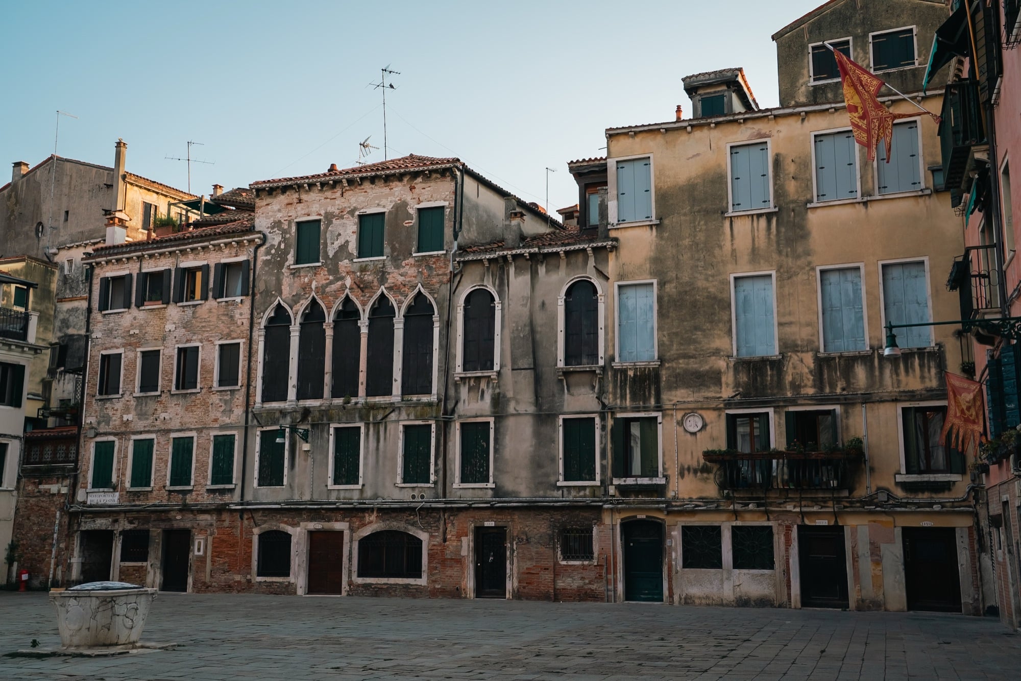 A quiet campo at dawn with a carved stone wellhead, weathered Gothic palazzo facades and Venetian lion flags flying