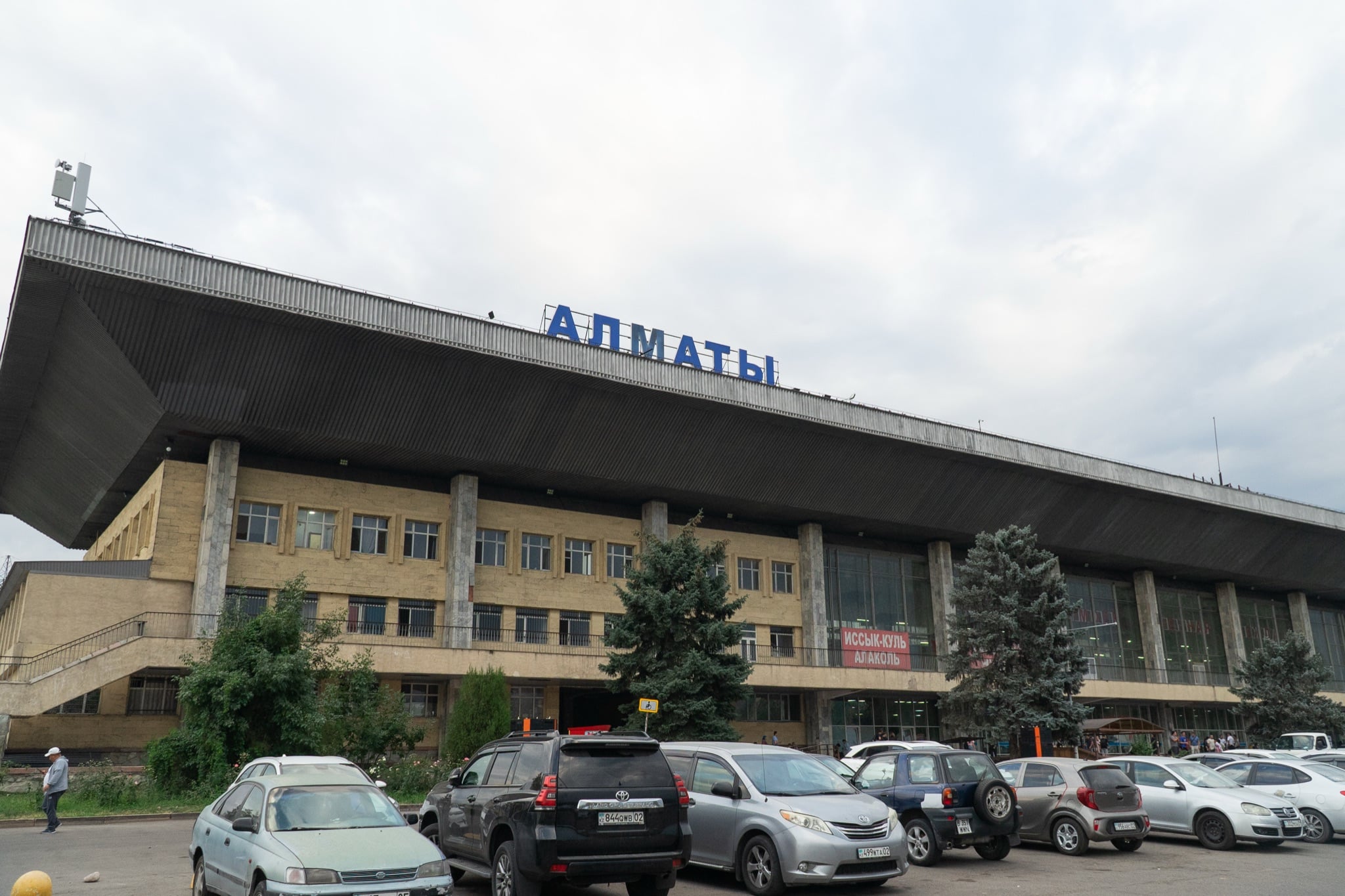 The modernist Almaty bus station beneath a grey sky, cars parked out front