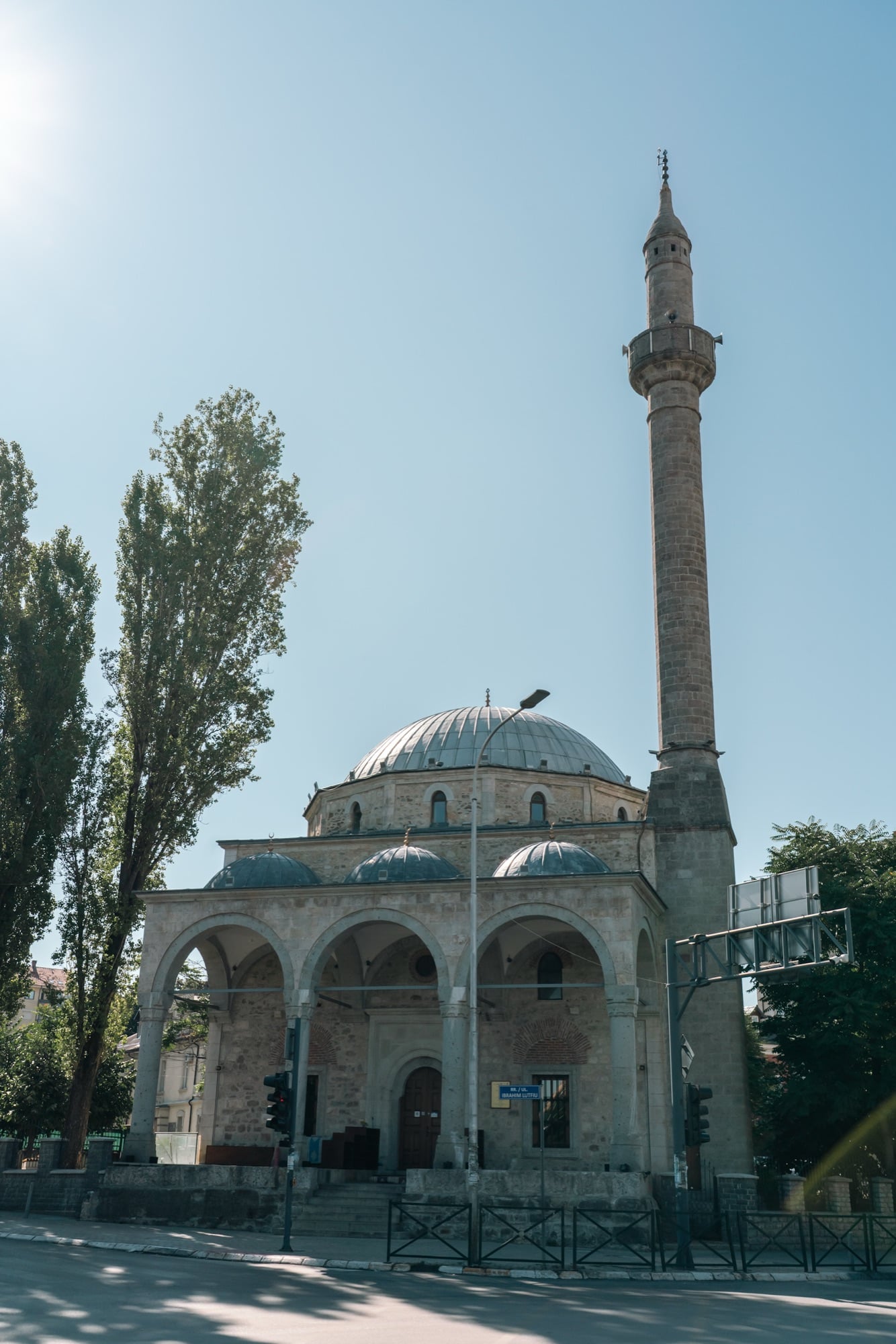 The lead domes and tall stone minaret of the Emperor's Mosque in Pristina framed by trees and a café awning