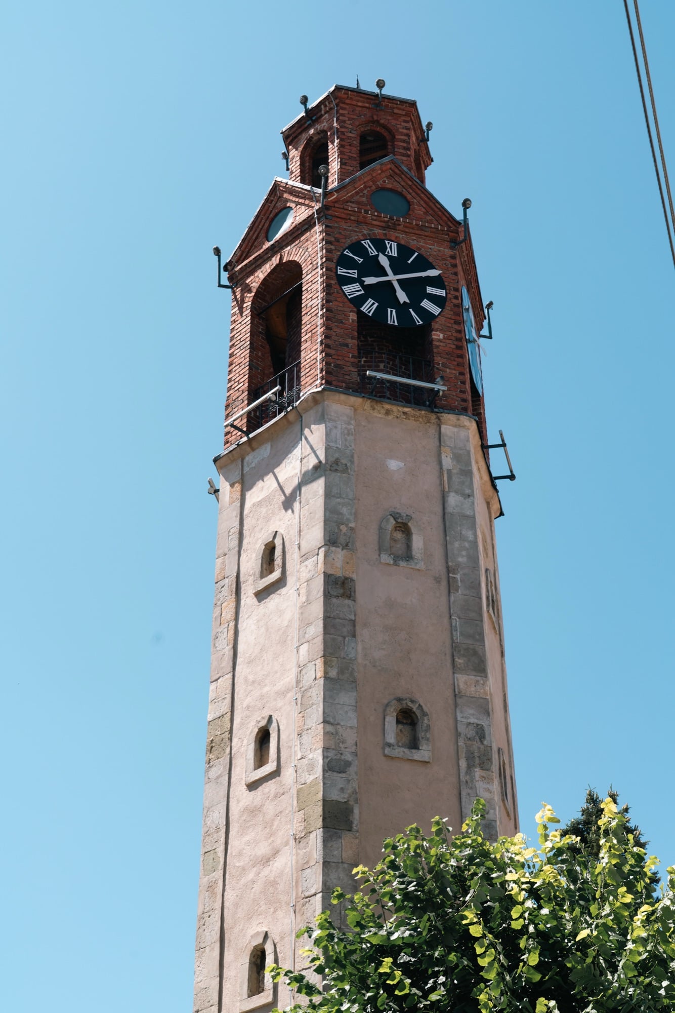The brick Clock Tower of Pristina with its black Roman-numeral face rising above a weathered stone base against a clear sky