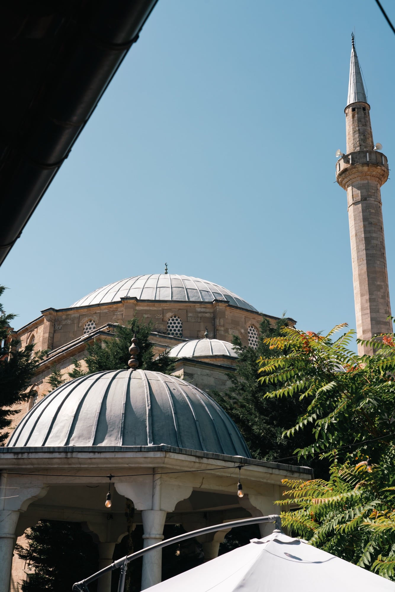 The stacked lead domes and stone minaret of an Ottoman mosque in Pristina seen past a café awning and greenery