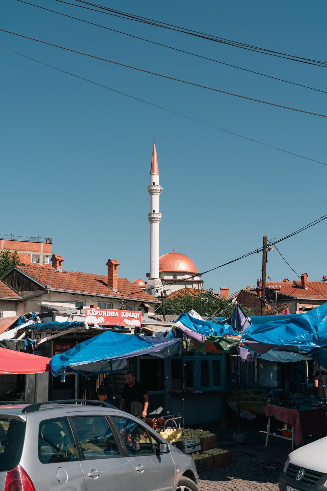 A white minaret and copper-domed mosque rising above the tarpaulins and produce stalls of a Pristina street market
