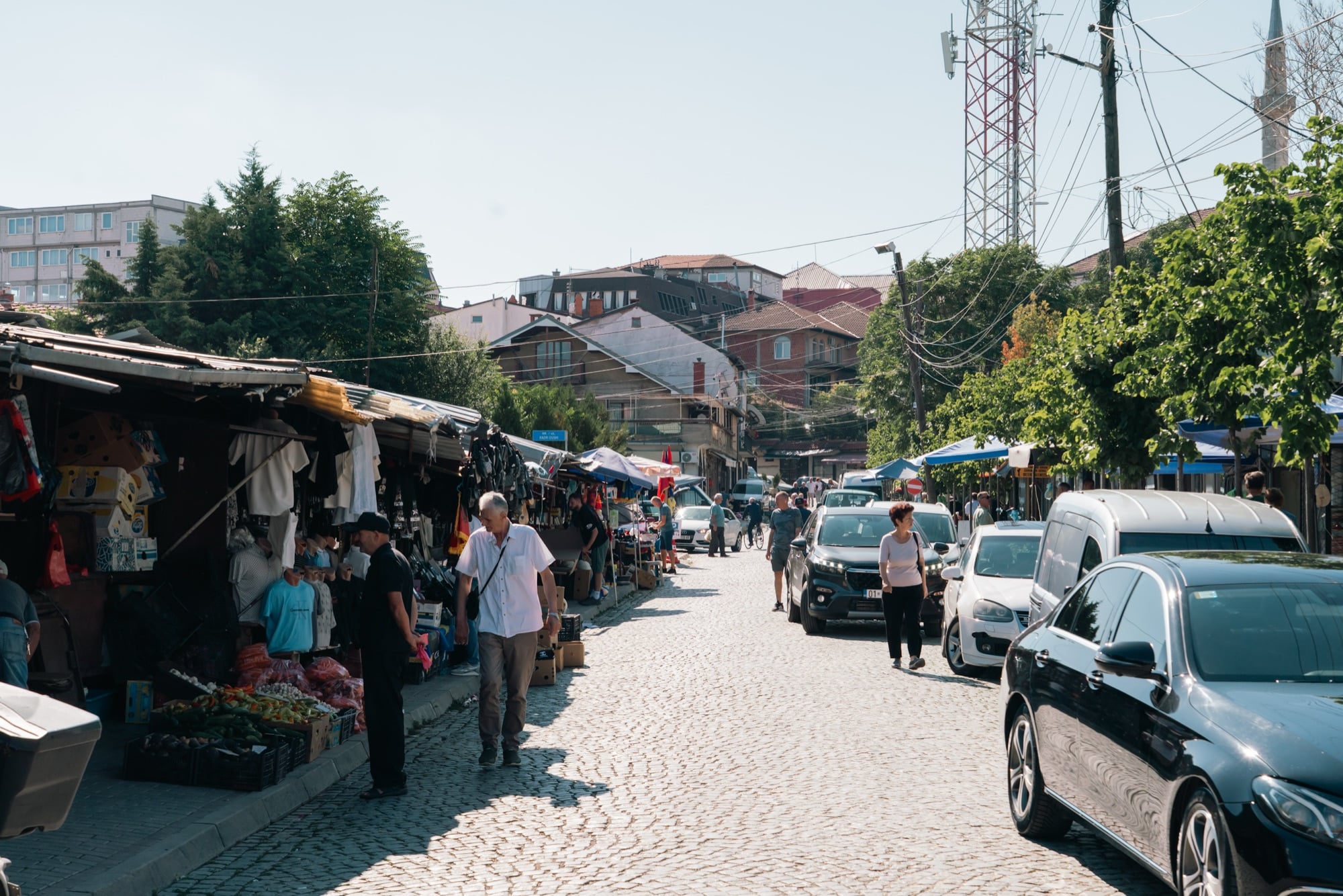Shoppers among the produce stalls of a Pristina market street, a telecoms mast and distant minaret rising above the rooftops