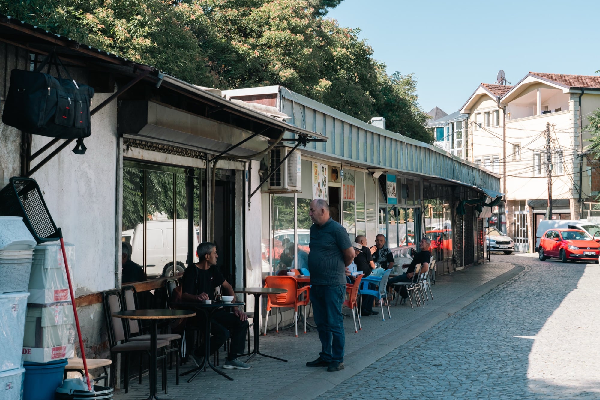 Men taking coffee at kiosk cafés along a cobbled Pristina side street, plastic chairs and stacked wares in the morning sun