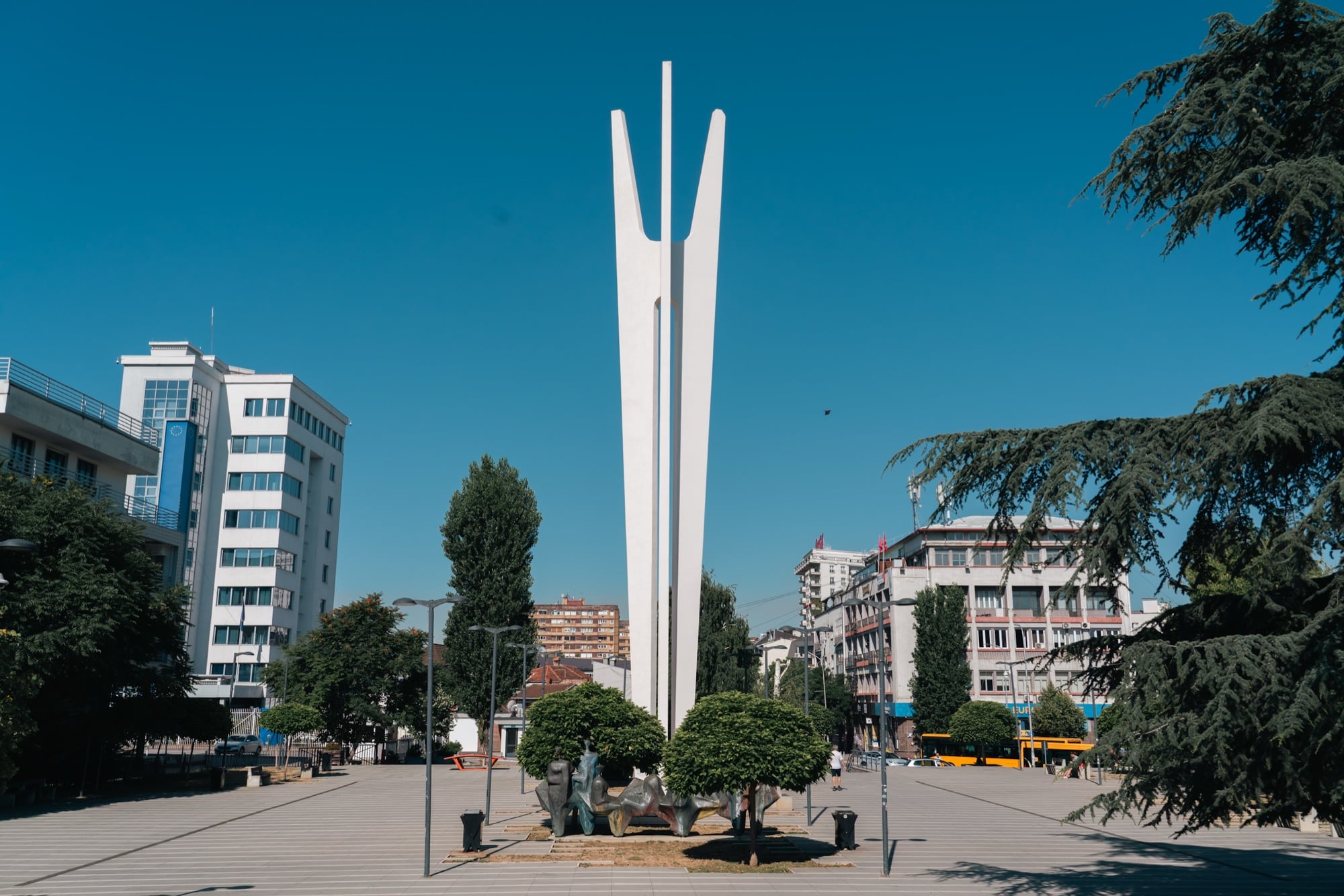 The tall white three-pronged Brotherhood and Unity Monument on a Pristina plaza, an EU-draped building alongside