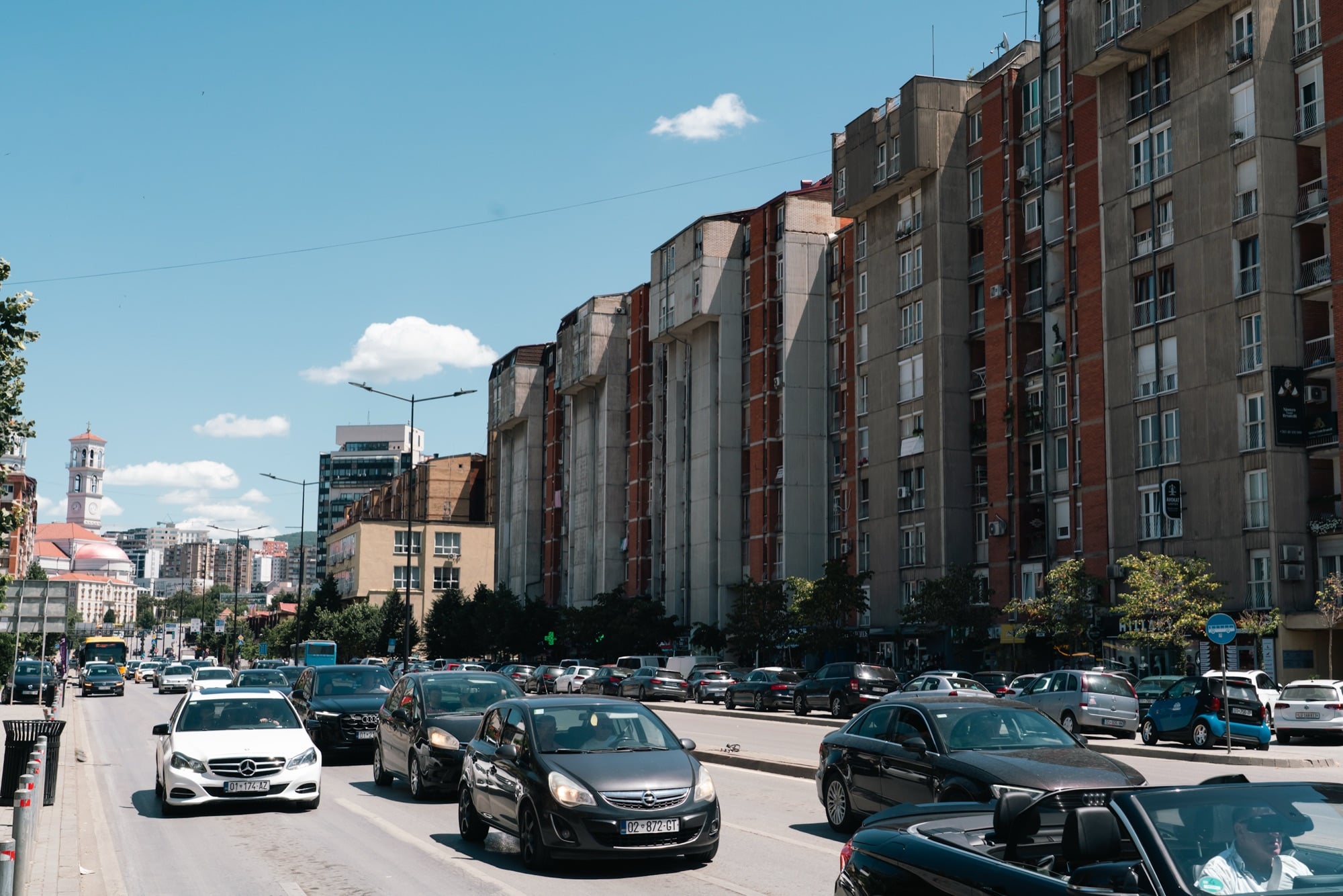 Traffic along a Pristina boulevard past socialist-era apartment blocks, the cathedral bell tower on the skyline beyond