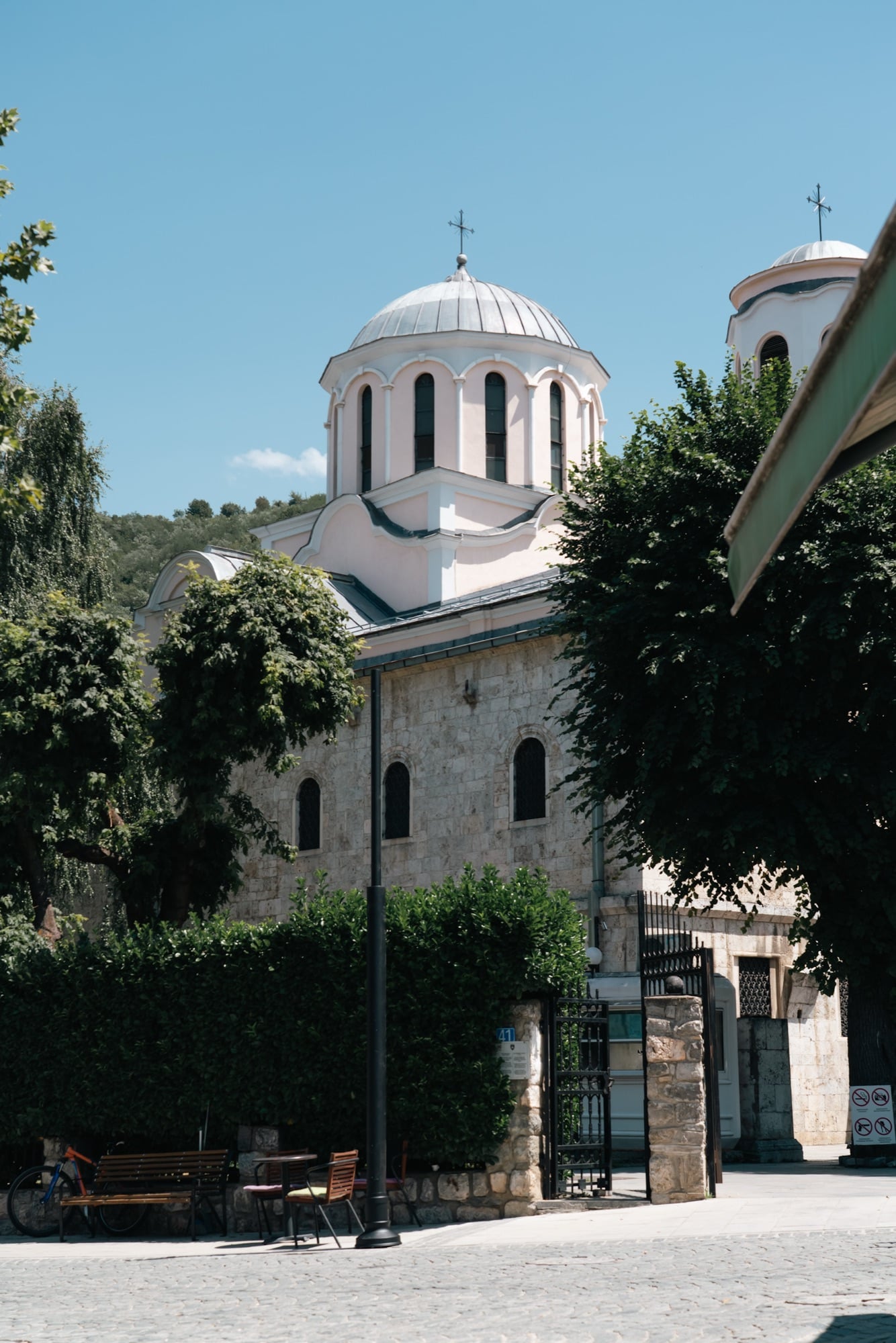 The domed St George's Orthodox Cathedral of Prizren rising behind trimmed hedges and shade trees, its cross catching the light