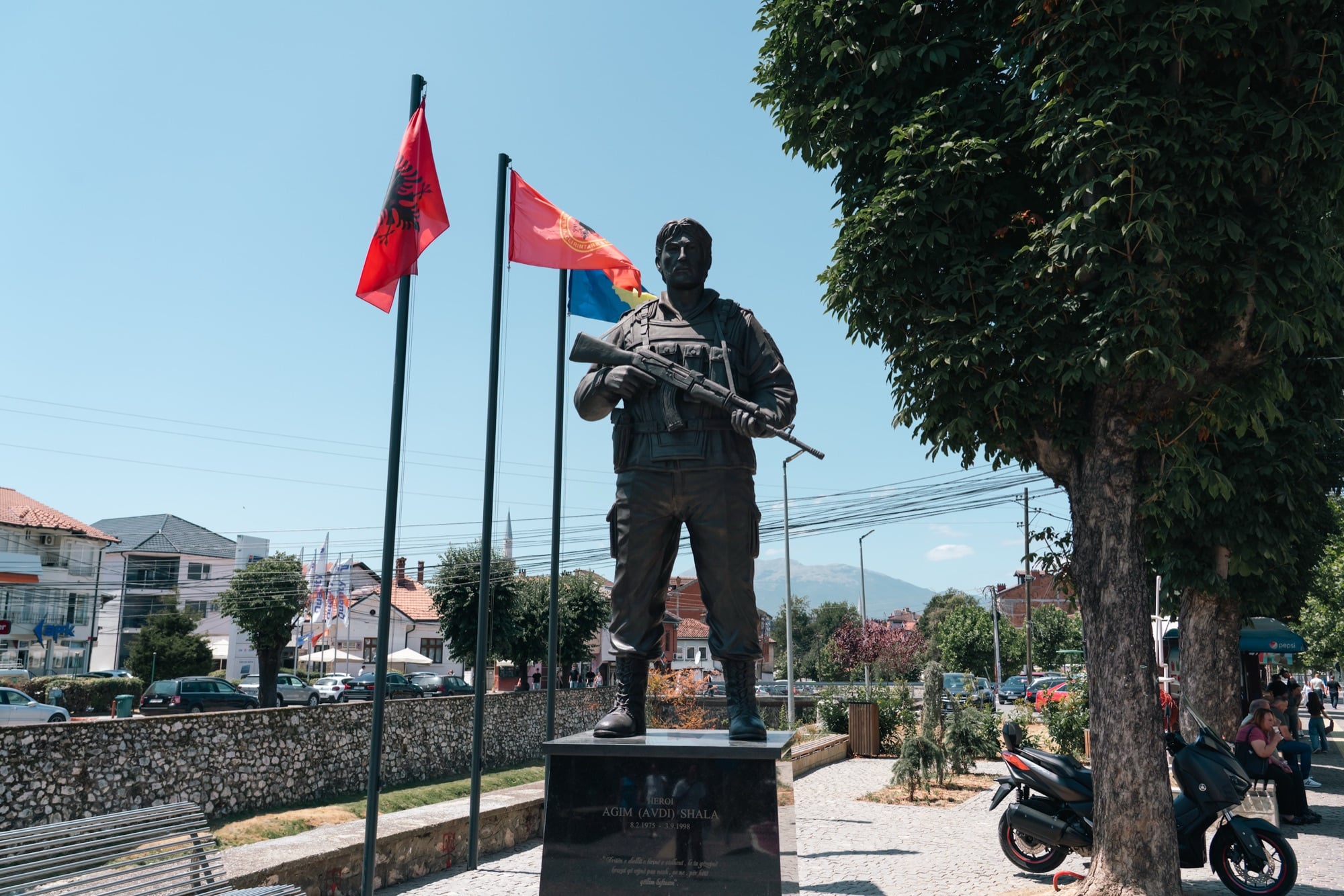 The bronze statue of KLA hero Agim Shala shouldering a rifle in a Prizren square, Albanian flags and mountains behind