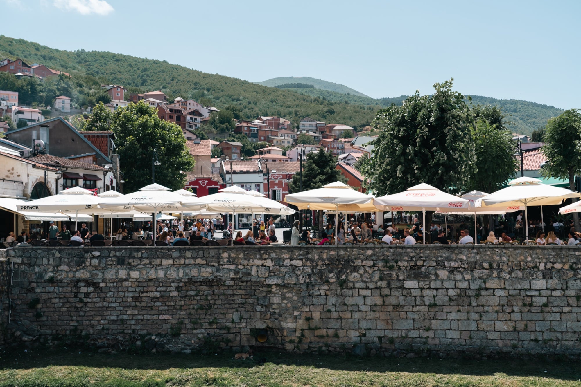 Prizren's riverside cafés packed under white umbrellas along the old stone embankment, tiled houses climbing the wooded hills beyond