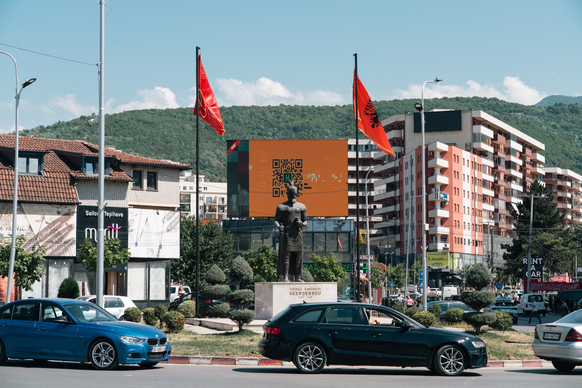 The Skanderbeg statue on a Prizren roundabout flanked by two red Albanian flags, apartment blocks and green hills beyond