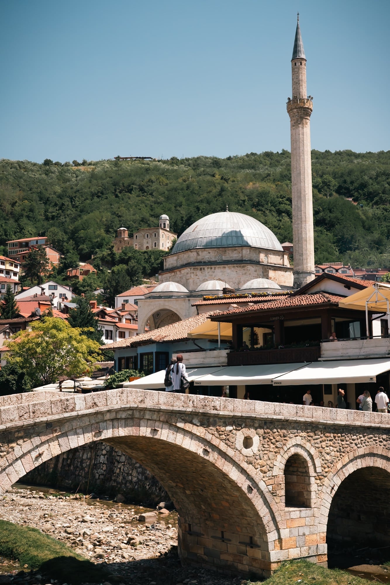 The lead dome and tall minaret of the Sinan Pasha Mosque rising above Prizren's old town, the Ottoman stone bridge crossing the Bistrica in the foreground
