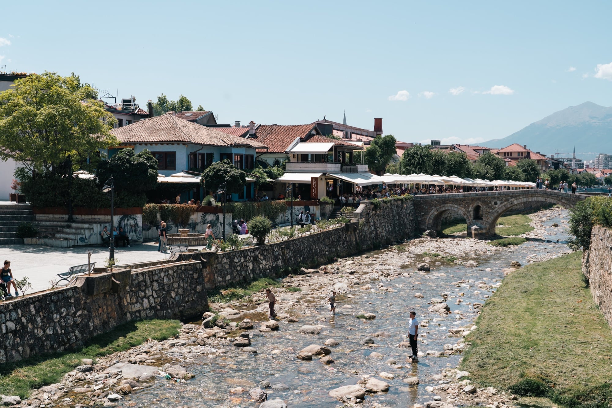 Cafés under white umbrellas lining the Bistrica river in Prizren, children wading in the shallow rocky stream below the old stone bridge