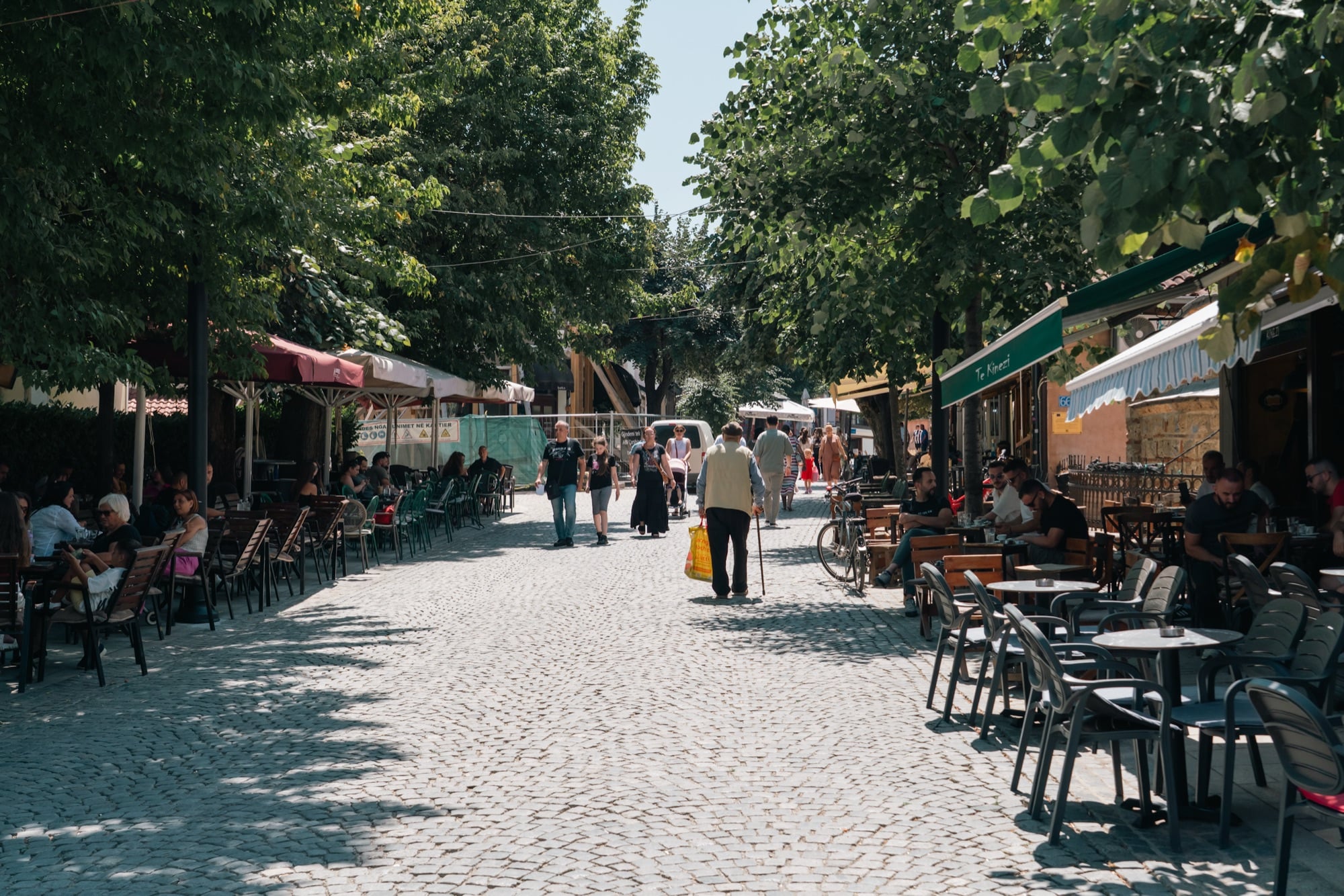 A tree-shaded cobbled café street in Prizren lined with tables and chairs, people strolling in the dappled light