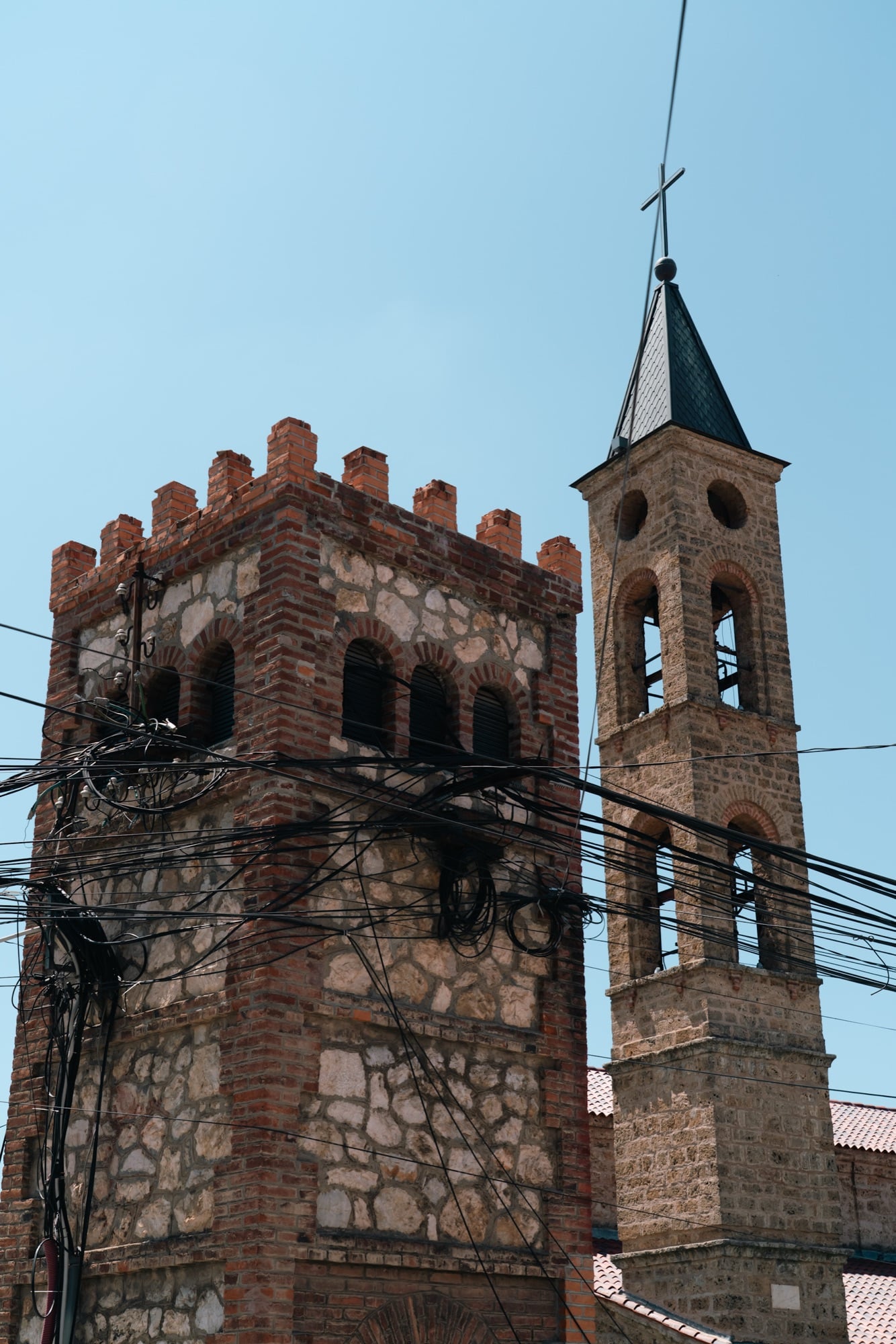 A crenellated brick-and-stone tower beside a bell tower topped with a cross in Prizren, tangles of overhead wires against the blue sky