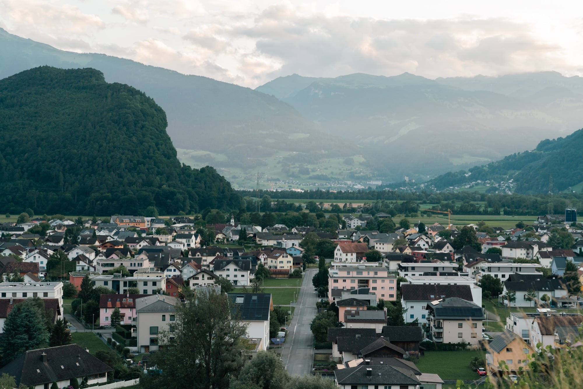 The village of Balzers spread across the valley floor at dusk, forested mountains fading into the evening haze