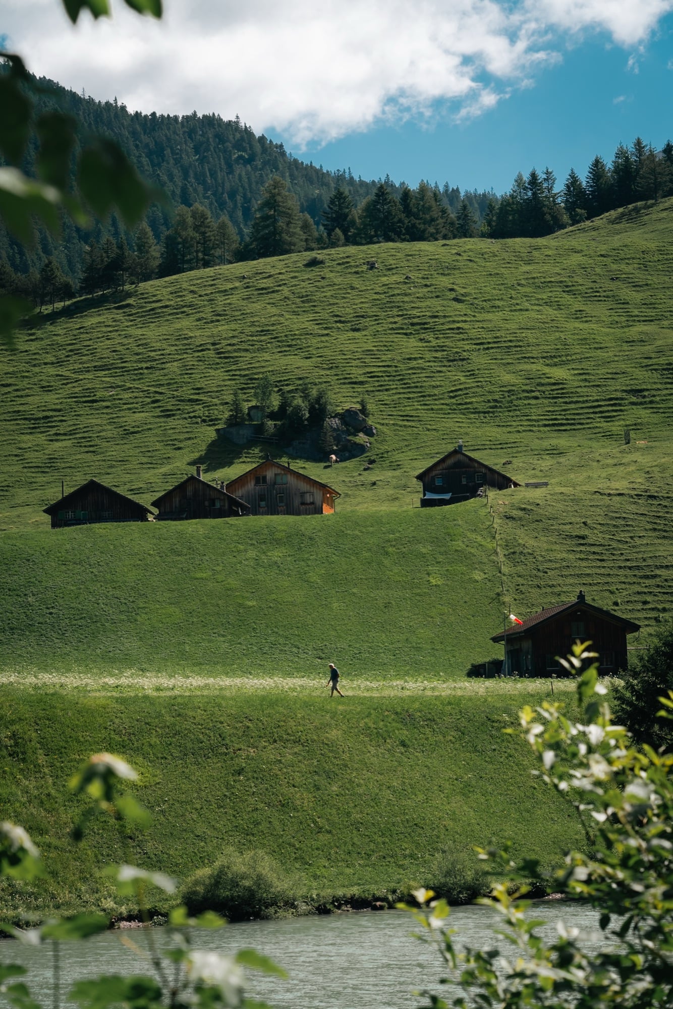 Dark-timber alpine huts scattered on a green slope above a turquoise river, a lone hiker on the path and a Liechtenstein flag flying