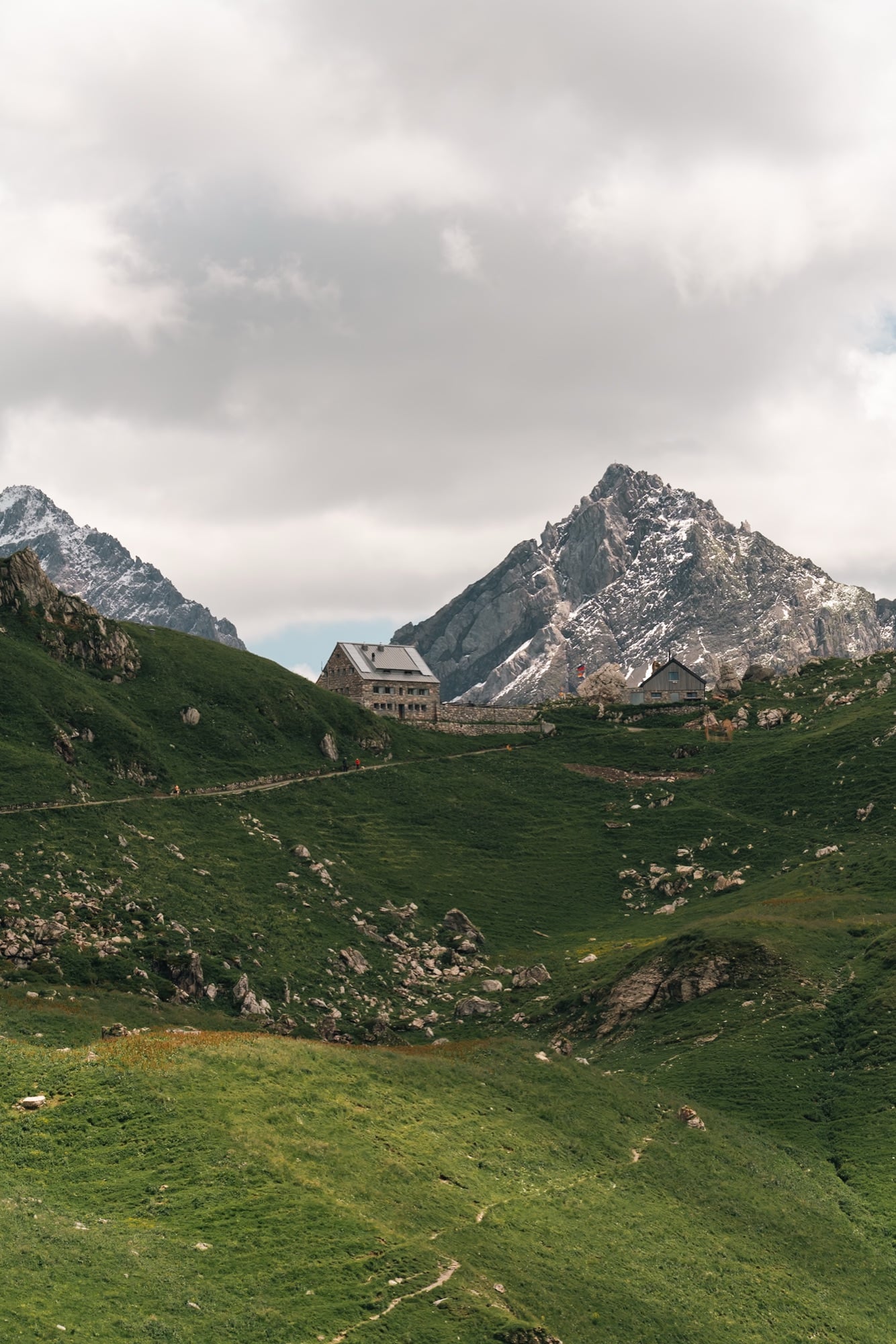 The stone Pfälzerhütte mountain hut on a green alp saddle below a jagged, snow-flecked peak, hiking trails threading the slopes