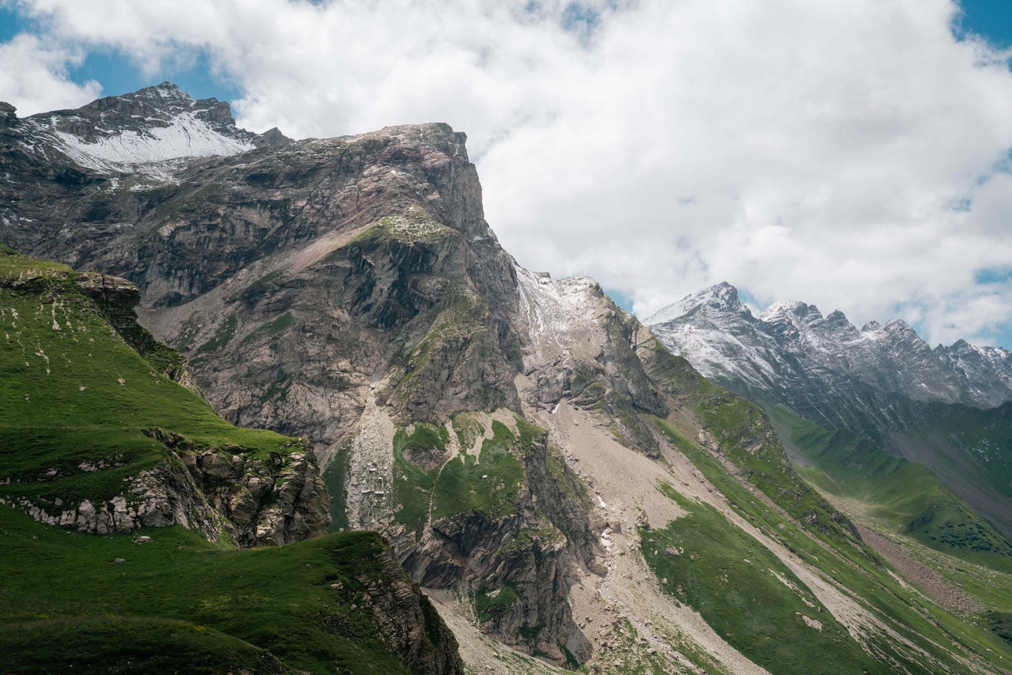 A rugged rock face streaked with snow rising above green alp slopes, the saw-toothed Rätikon ridgeline beyond