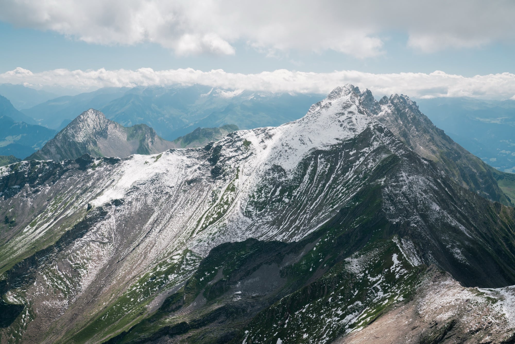 A high summit panorama of snow-patched ridgelines fading into layered blue ranges above a distant sea of cloud