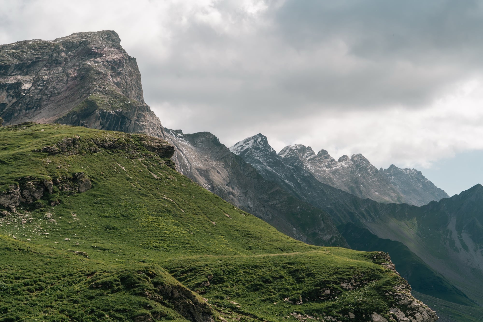 A green alp shoulder falling away below a saw-toothed rocky ridgeline under grey cloud