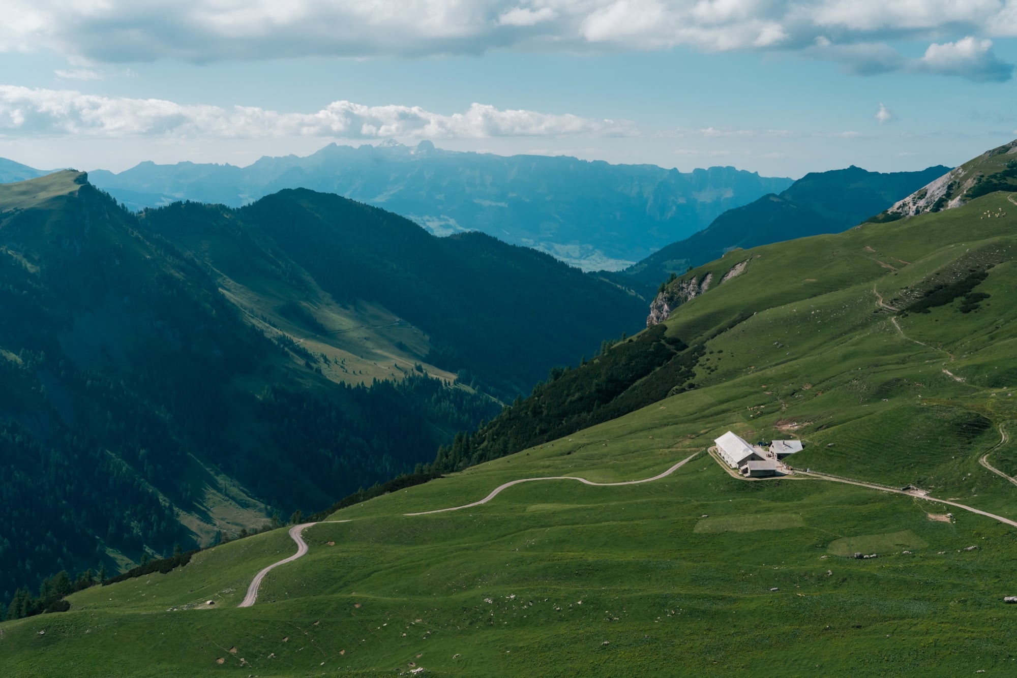 A sweeping view over green high pastures and a lone alp dairy, layered blue ridges receding to the valley beyond