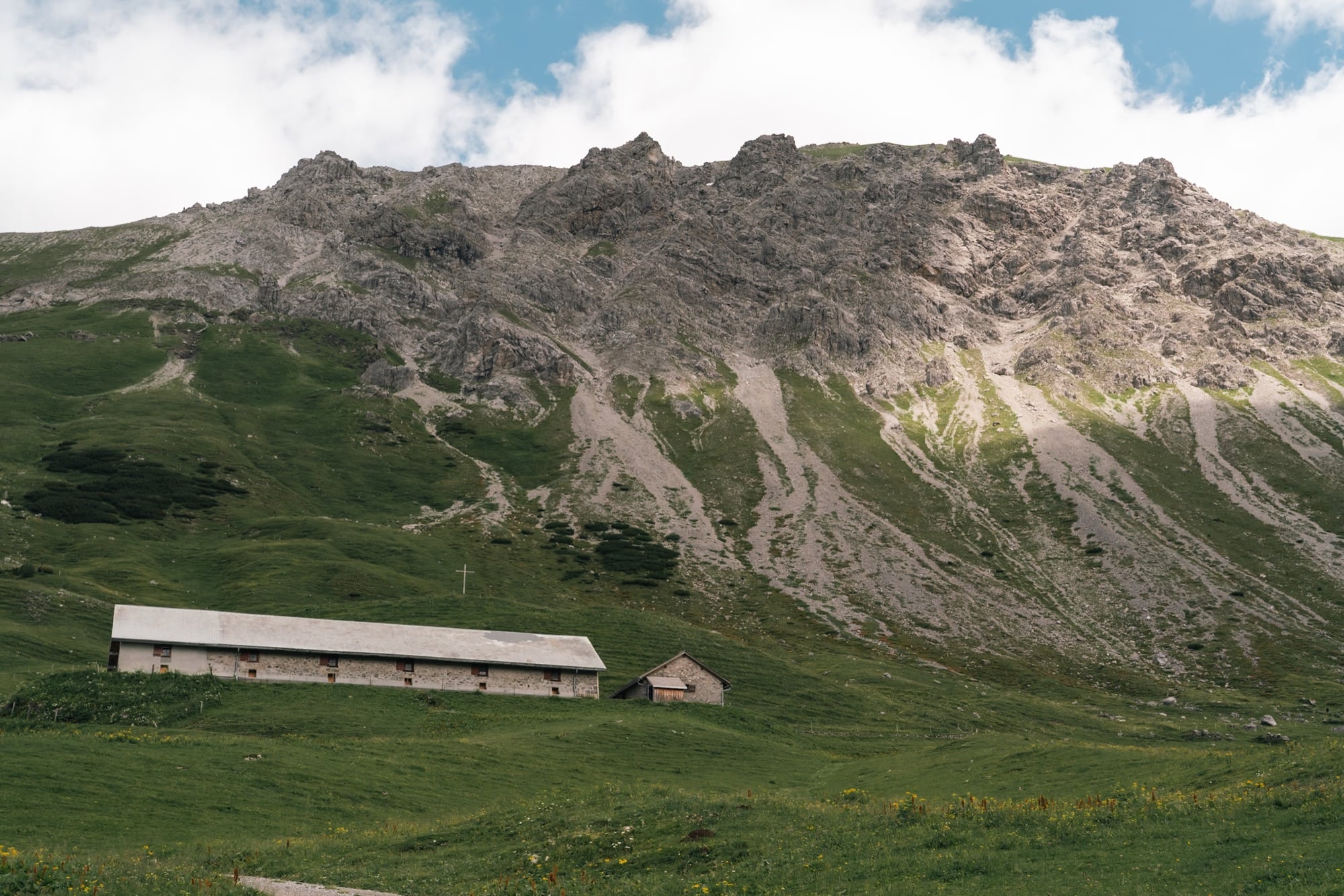 A long low stone alp barn on a green shelf below a scree-streaked ridge, a summit cross on the slope above