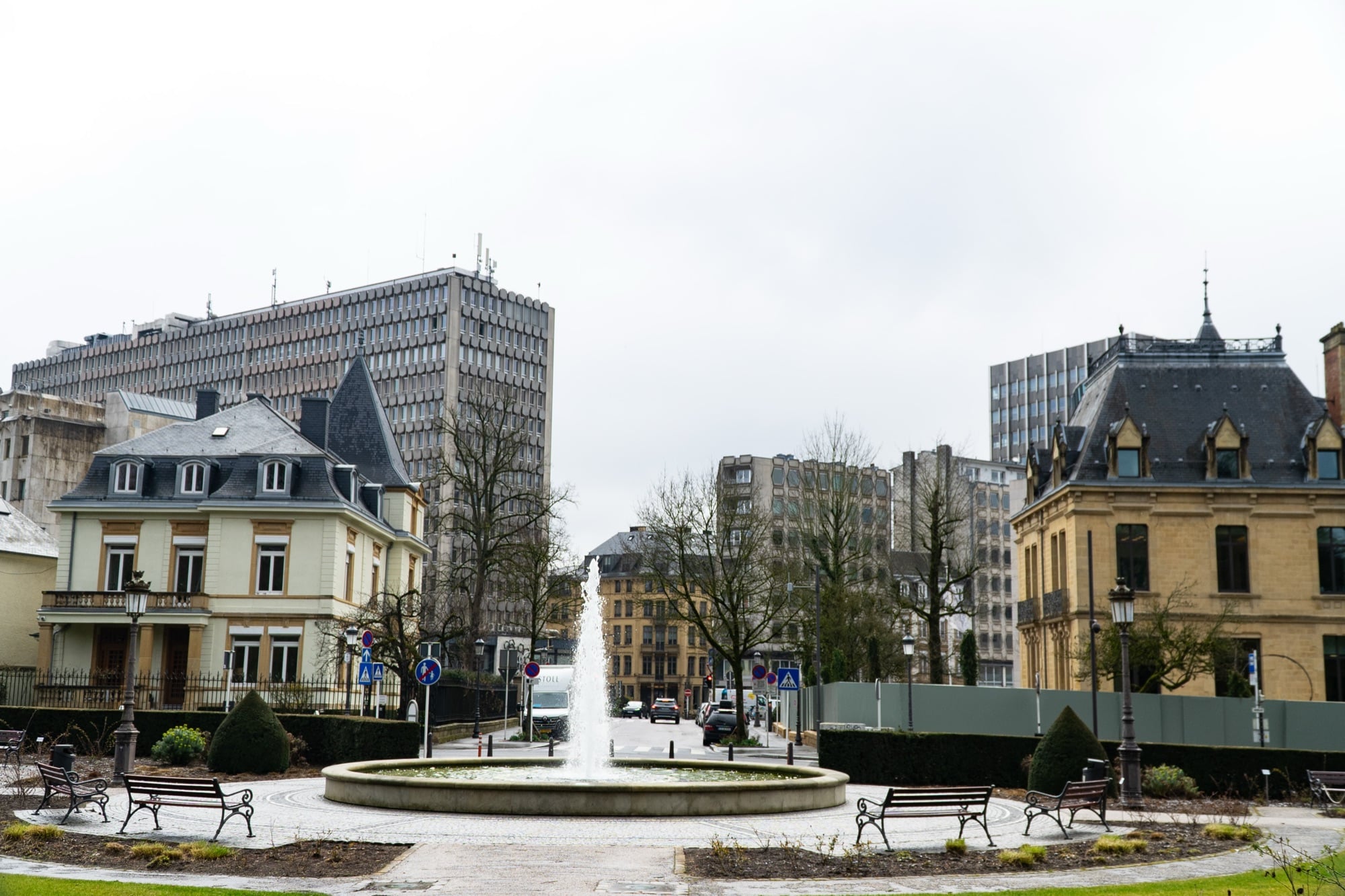 A fountain playing in a garden square ringed with benches, city buildings behind, Luxembourg City