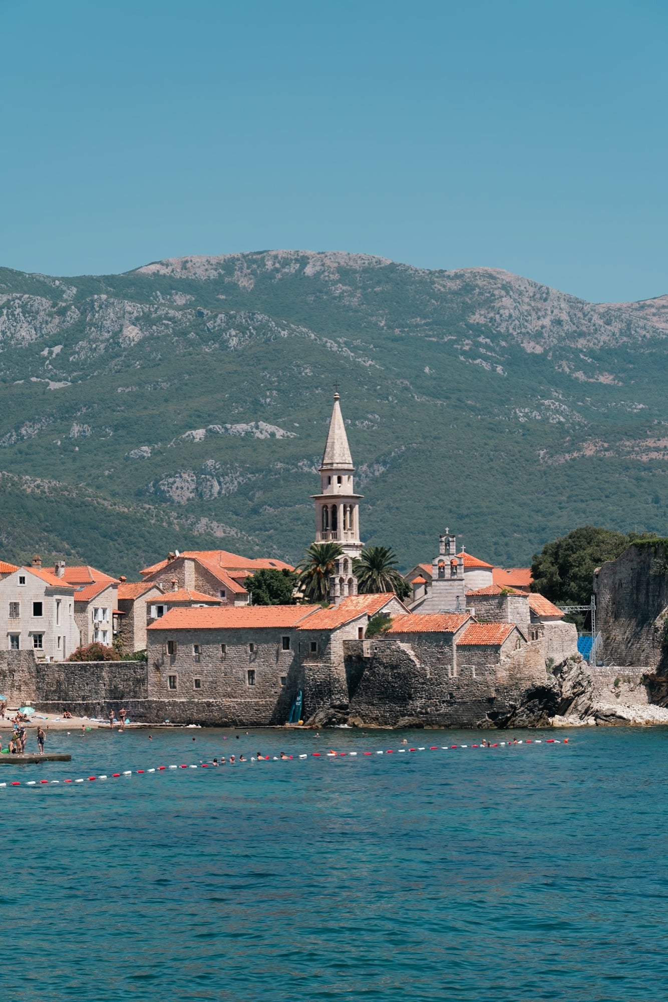 Budva's walled old town seen across the sea, the stone bell tower of its church rising above terracotta roofs beneath a bare mountain