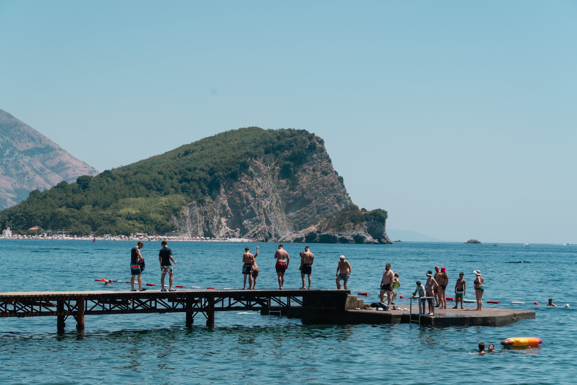 Swimmers gathered on a wooden jetty off a Budva beach, the green headland of Sveti Nikola island rising across the bay