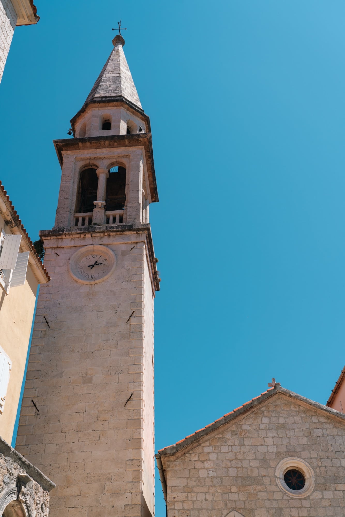 Looking up at the tall stone bell tower and clock of a church in Budva's old town against a deep blue sky