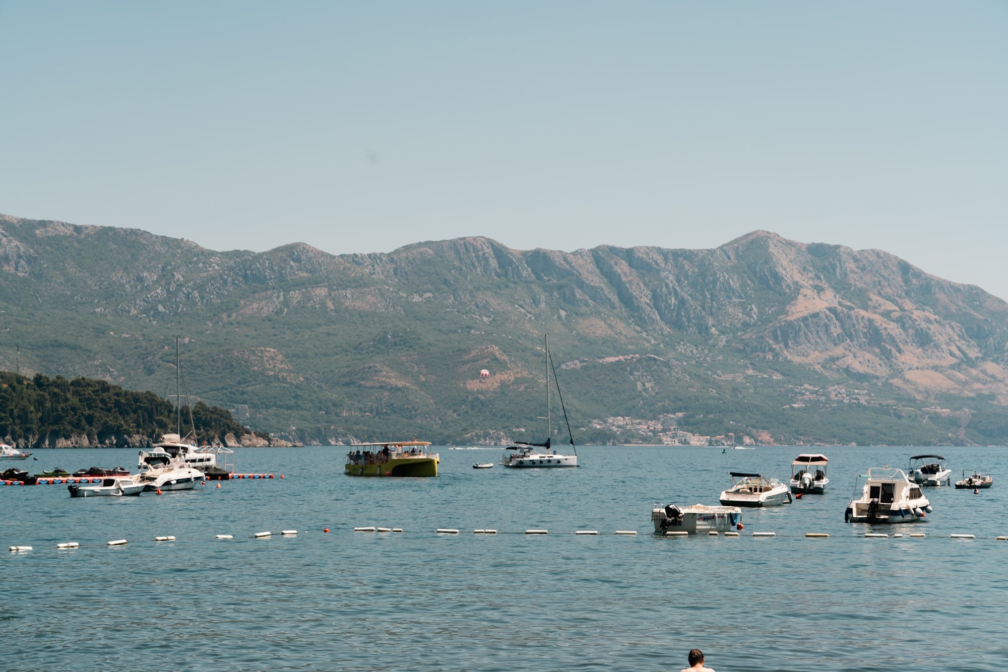 Pleasure boats moored inside a swim line off Budva, a parasail drifting over the long mountain ridge across the bay