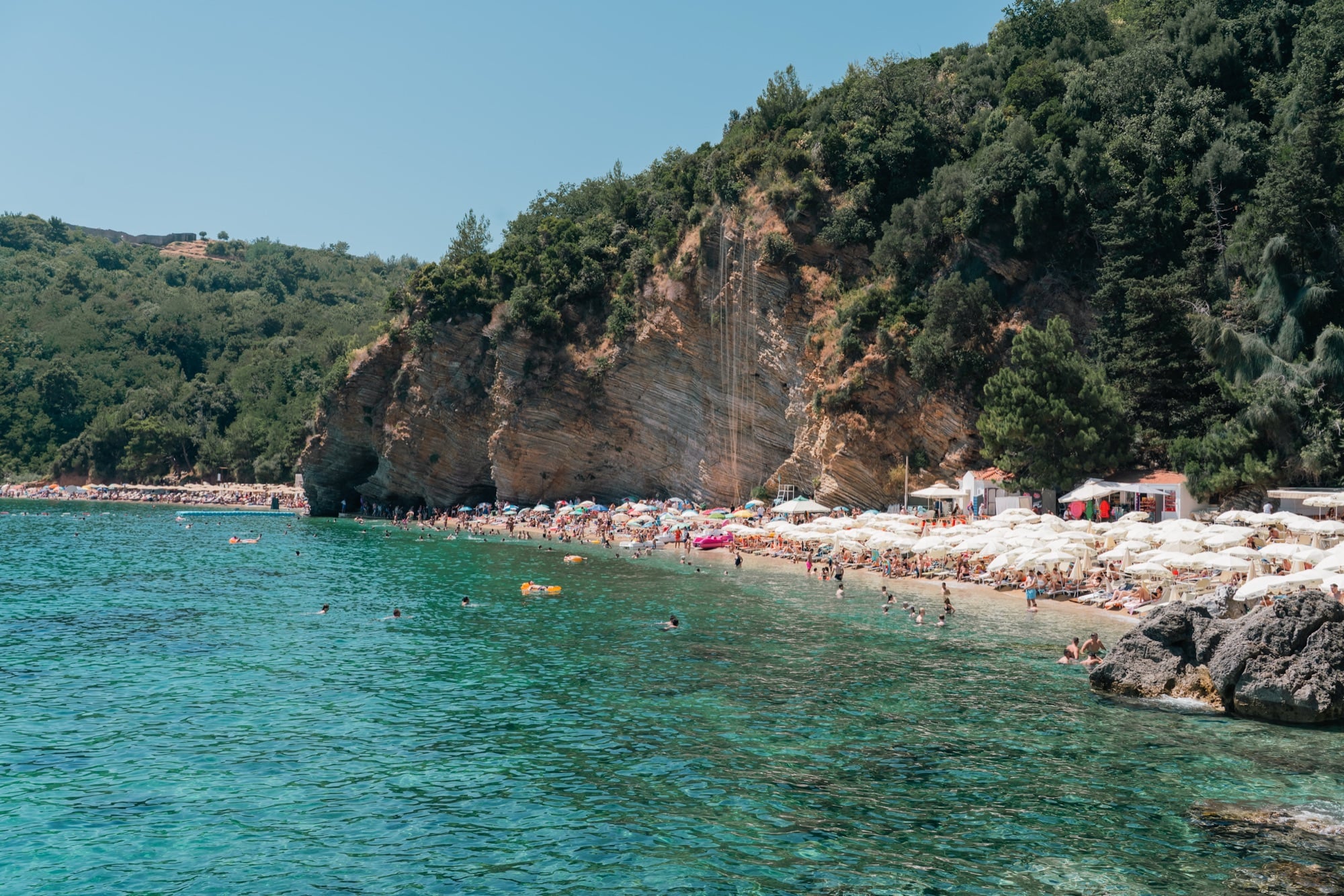 A crowded pebble cove near Budva backed by a striped ochre cliff, sun umbrellas lining the shore and swimmers in the turquoise shallows