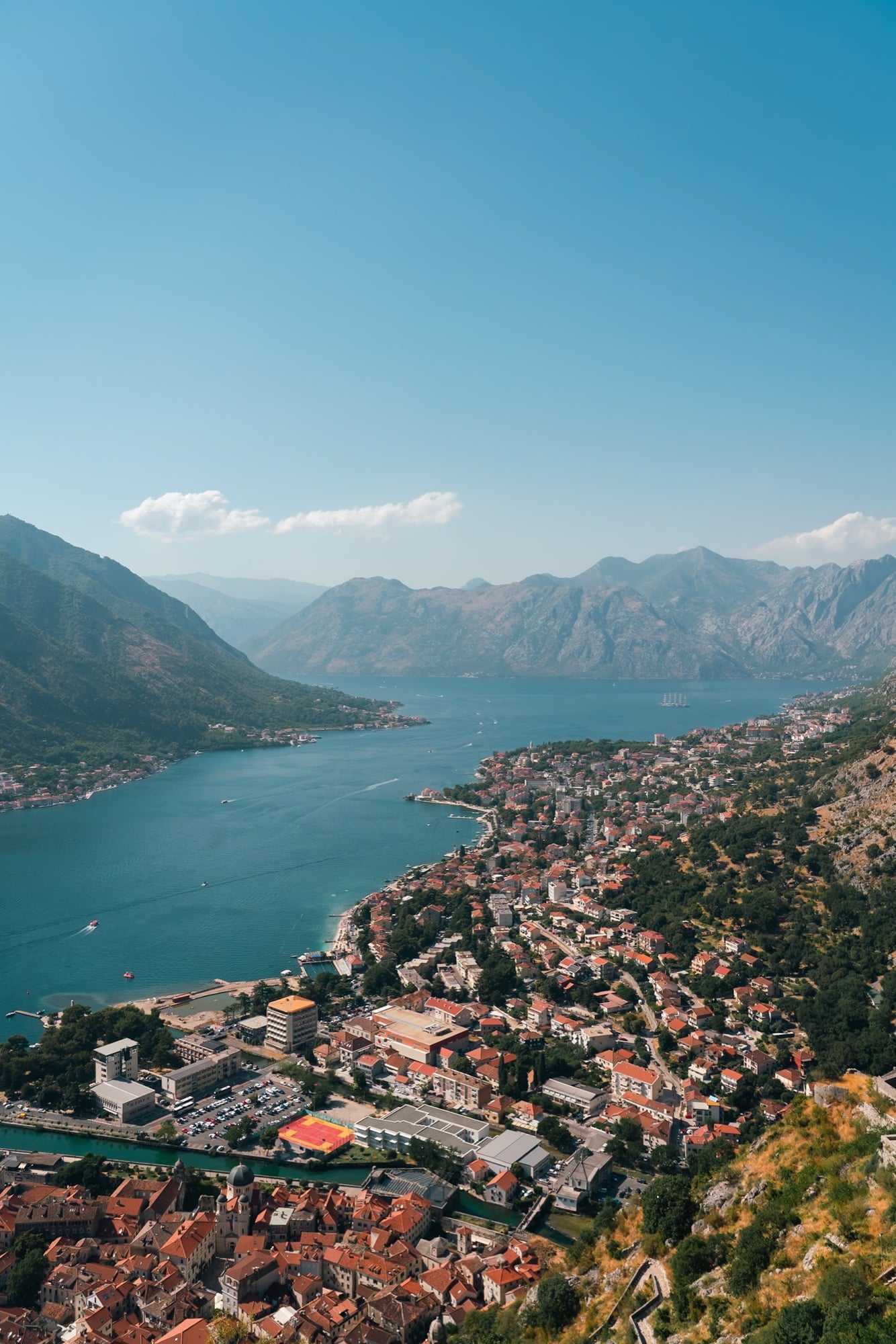 An aerial view from the fortress trail looking down over Kotor's red-roofed old town, the fjord-like bay winding between steep grey mountains beyond