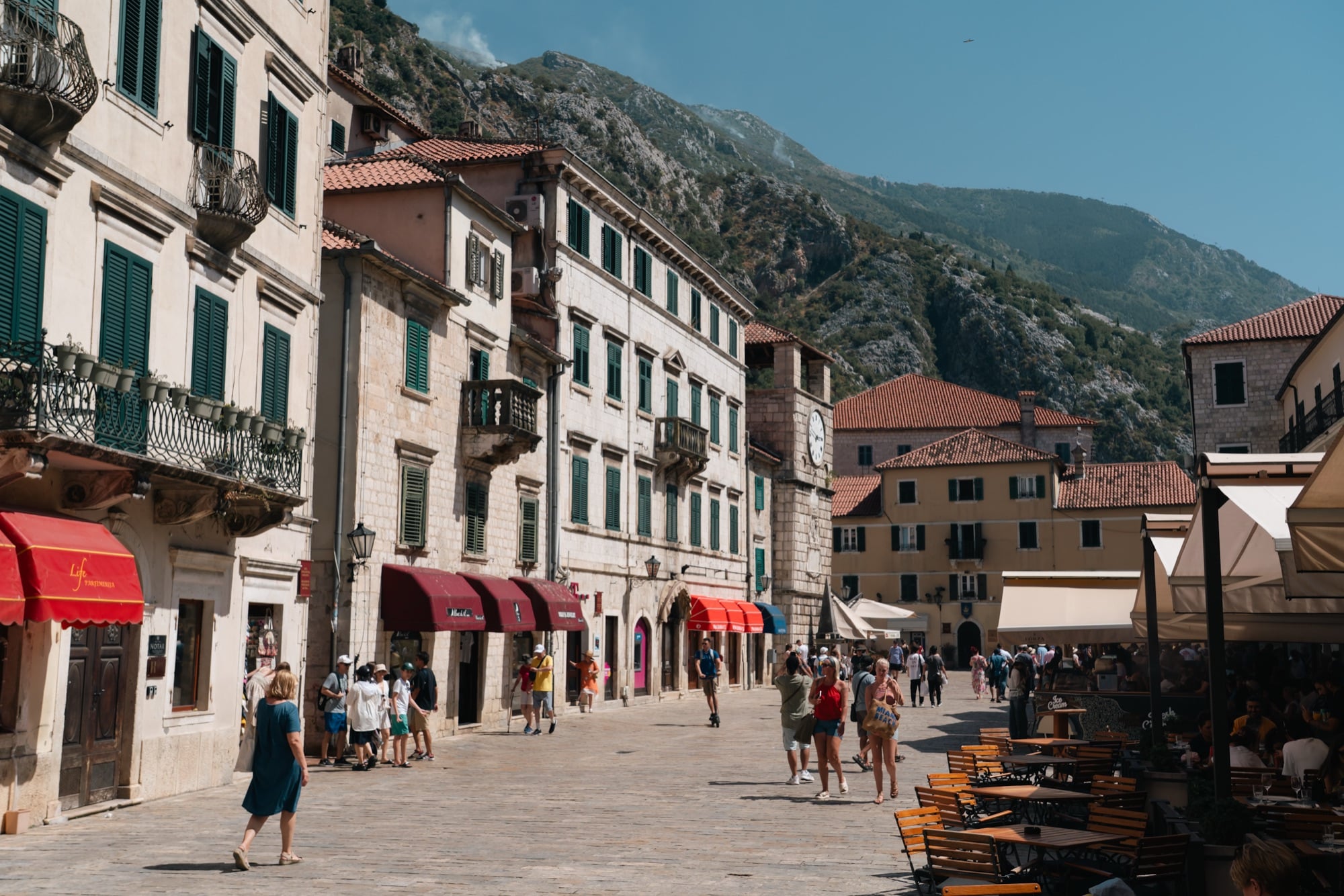 A sunlit square in Kotor's old town with café tables and the medieval clock tower, stone façades set below a steep mountainside