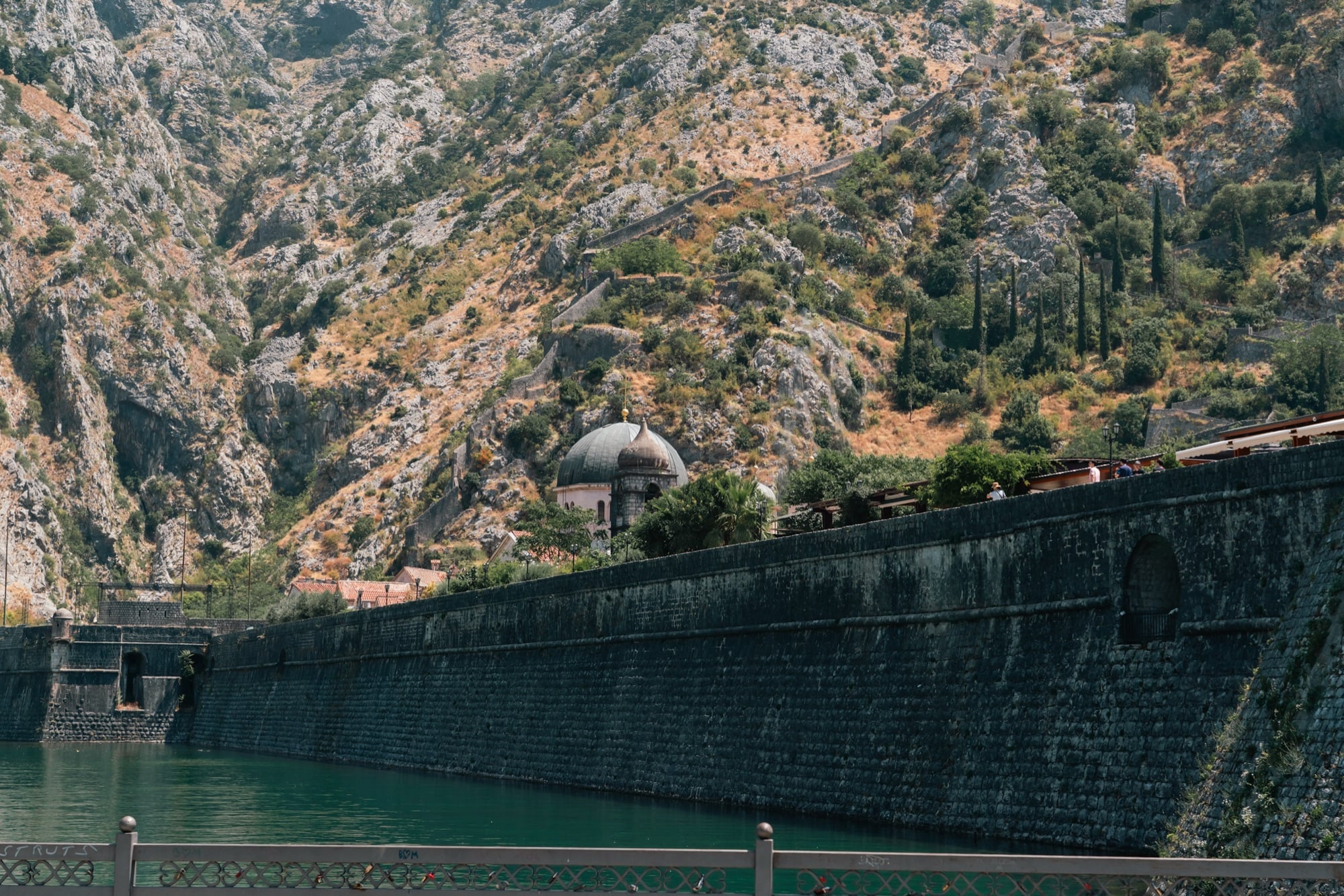 Kotor's massive stone rampart running beside the green river channel, a domed church tucked against the bare mountainside above
