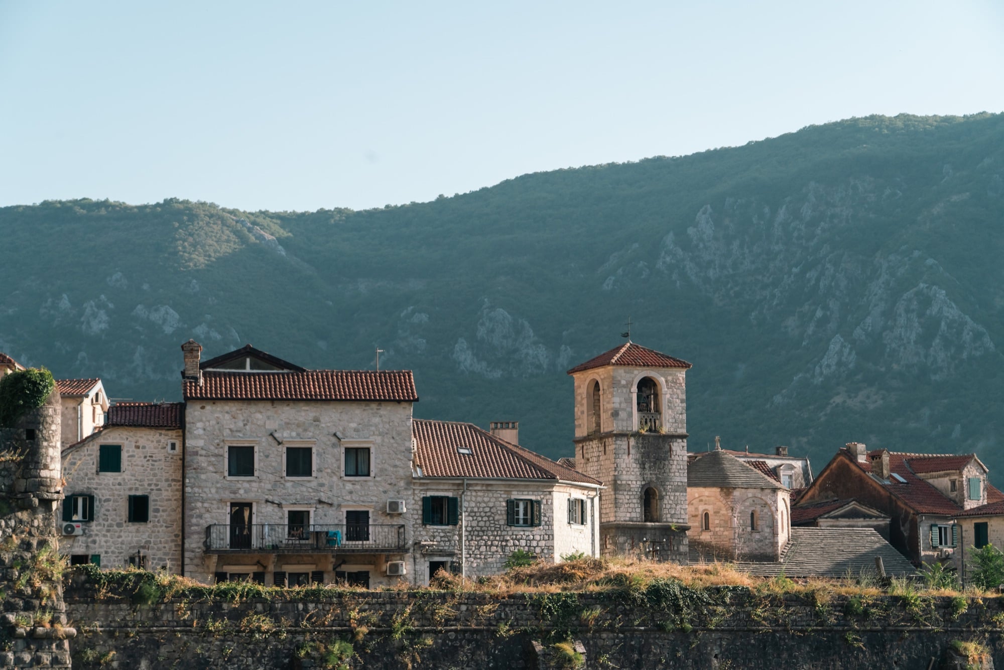 Old stone houses and a slender bell tower of Kotor catching the early light beneath a wooded mountain ridge