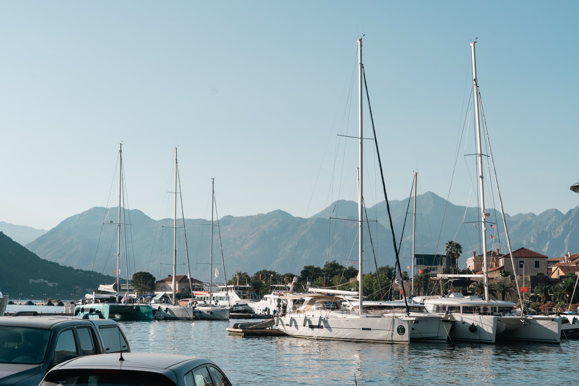 Sailboats and yachts moored in Kotor's marina, their masts standing against the hazy layered mountains across the bay