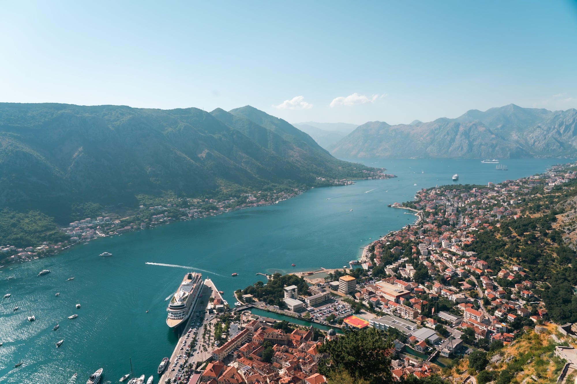 A high panorama of the Bay of Kotor with a cruise ship berthed at the waterfront, the town spread along the shore beneath towering mountains