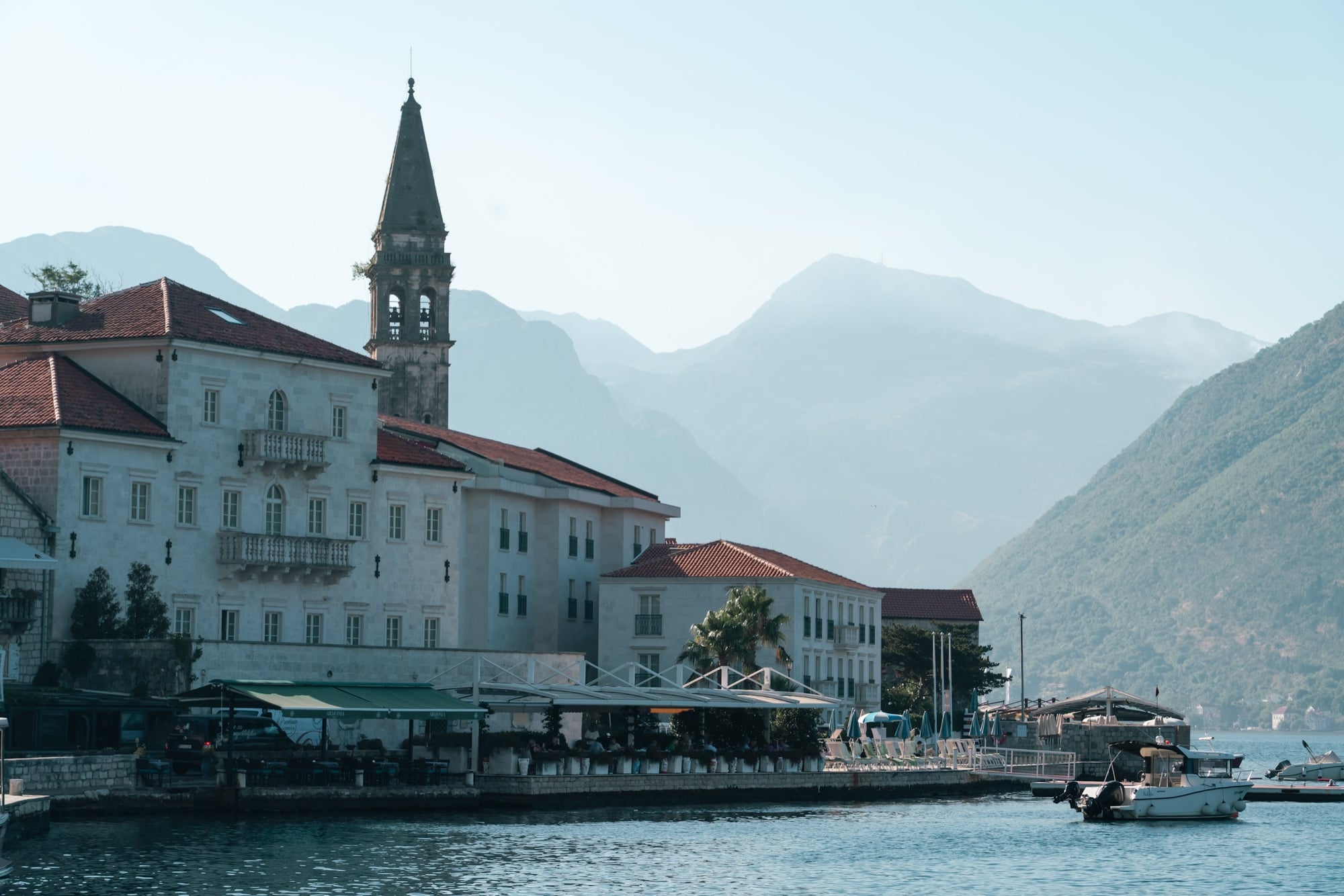 Perast's waterfront of Baroque stone palazzi and its towering bell tower, hazy mountains folding into the distance beyond the calm bay