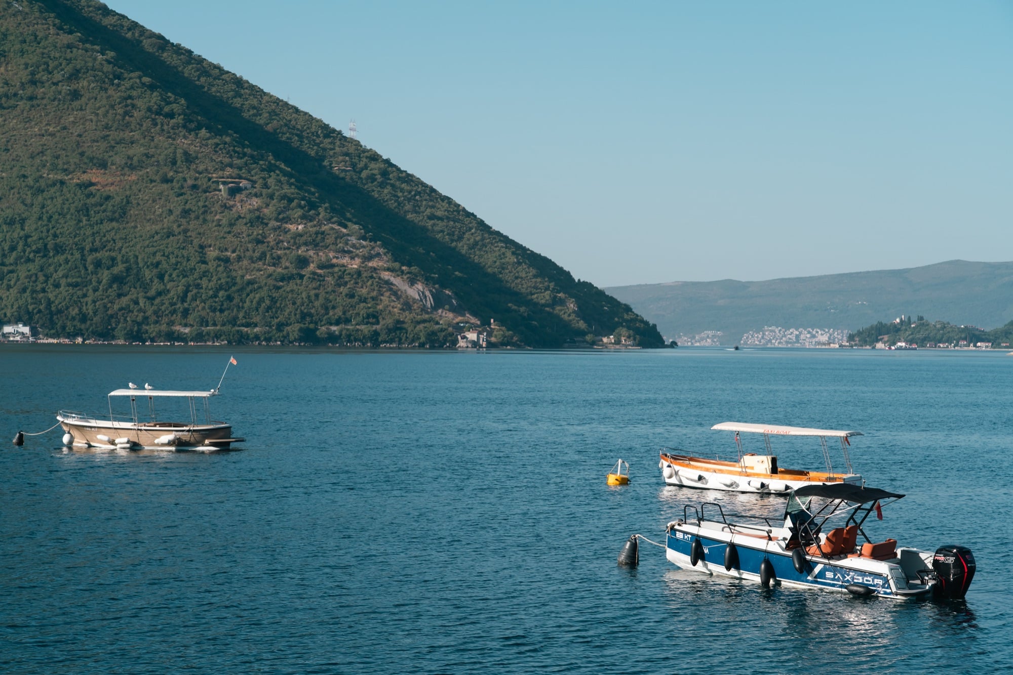 Small boats moored on the glassy Bay of Kotor off Perast, a green mountain ridge plunging straight to the water behind them