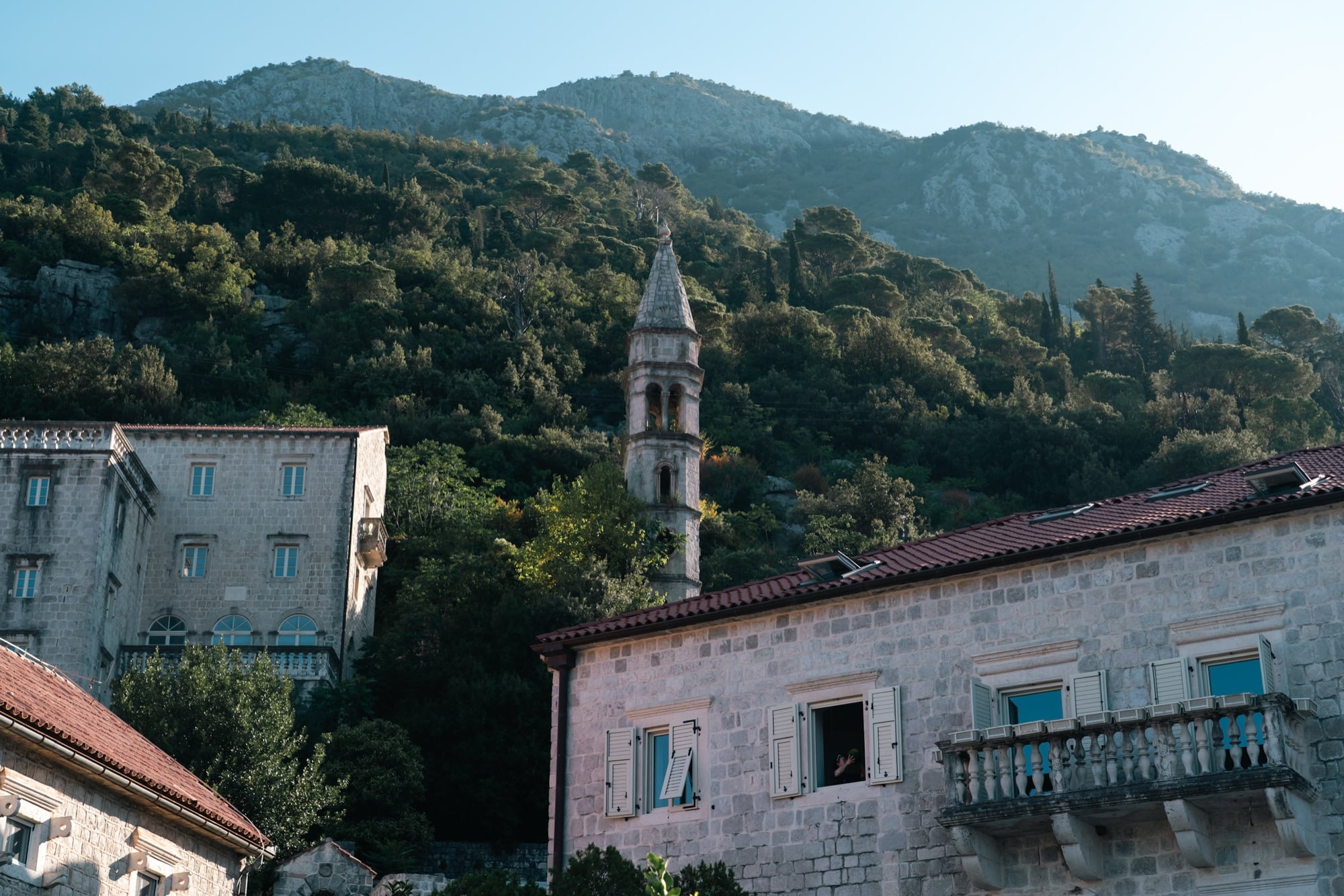 Sunlit stone houses of Perast climbing the hillside beneath a slender bell tower and rugged mountains