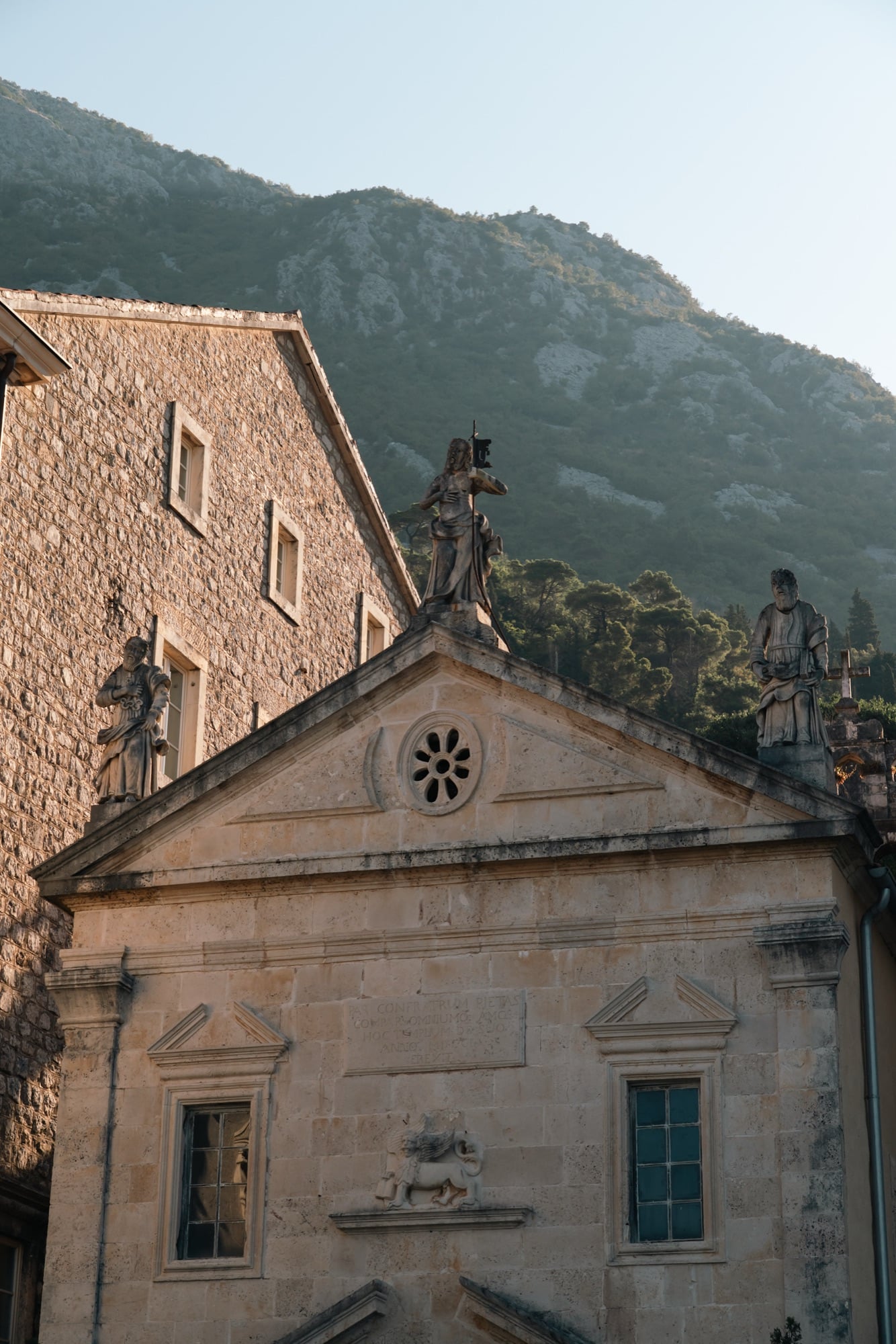 The Baroque stone façade of a Perast church topped with saintly statues, a carved Venetian lion above the door and mountains rising behind