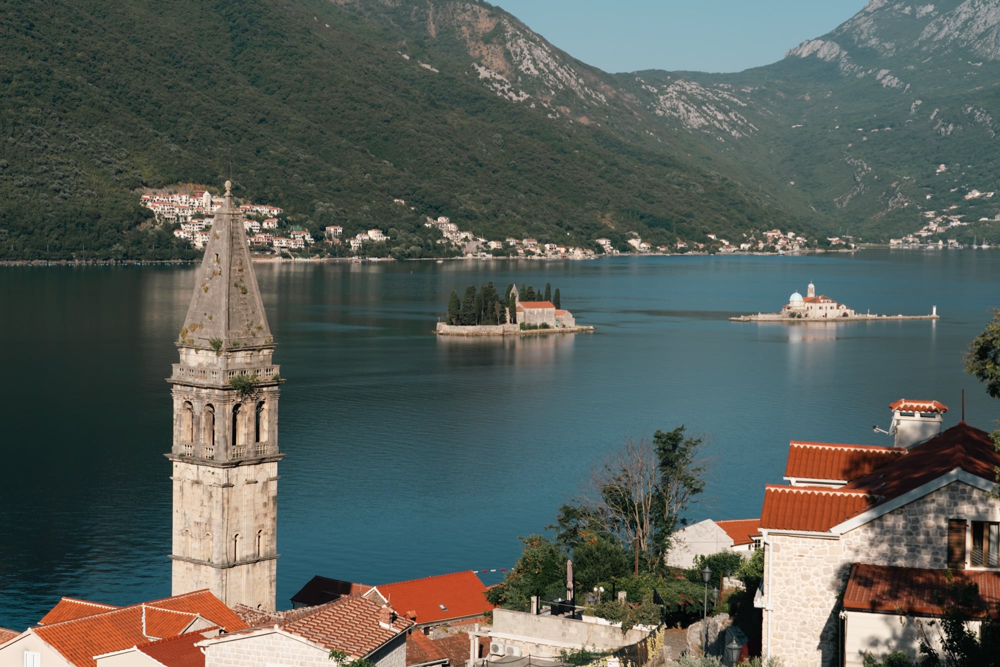 Perast's bell tower in the foreground with both islets — St George and Our Lady of the Rocks — set out on the deep-blue bay beyond