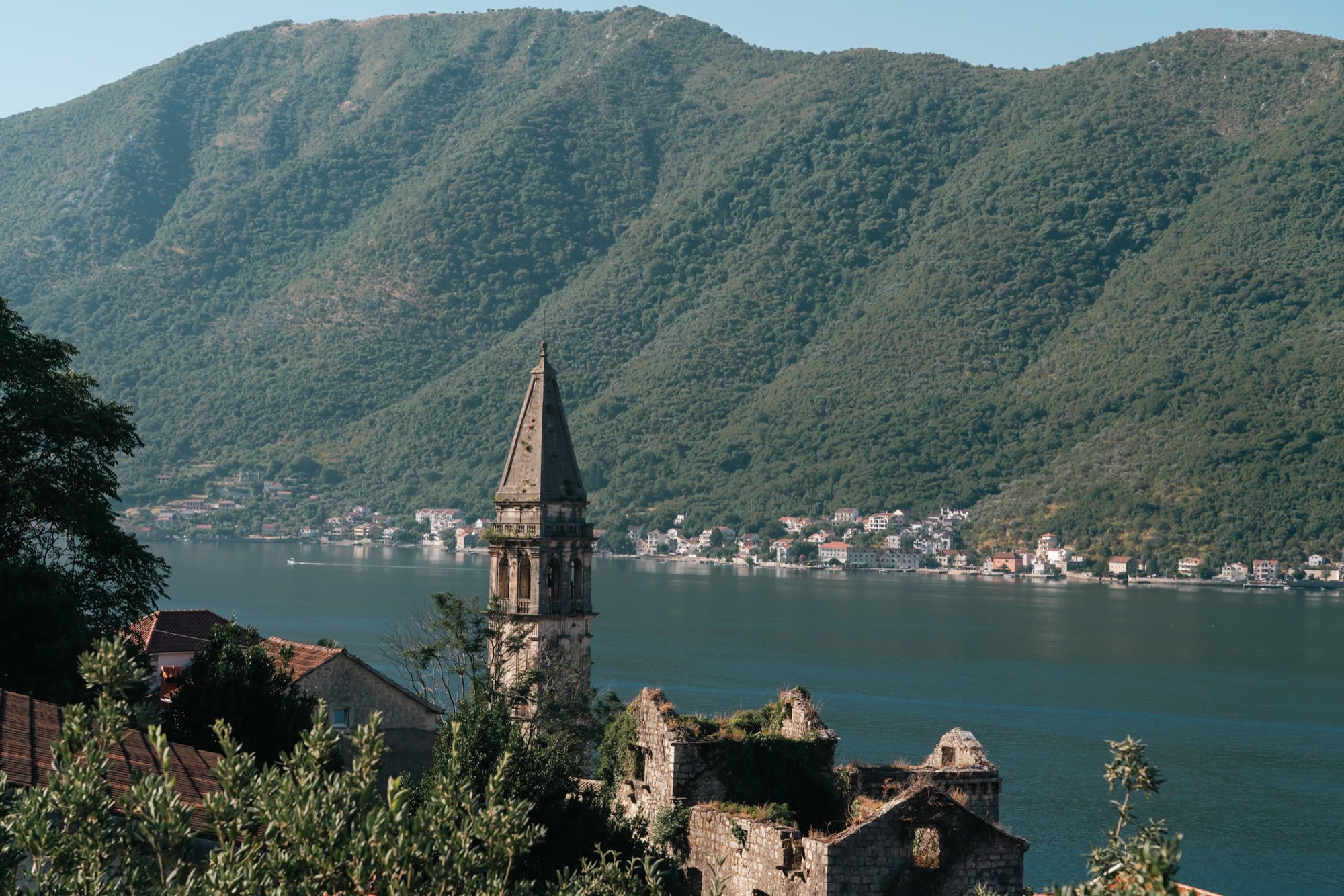 The lone stone bell tower of Perast standing tall above the water, a village and forested mountains lining the far shore of the bay