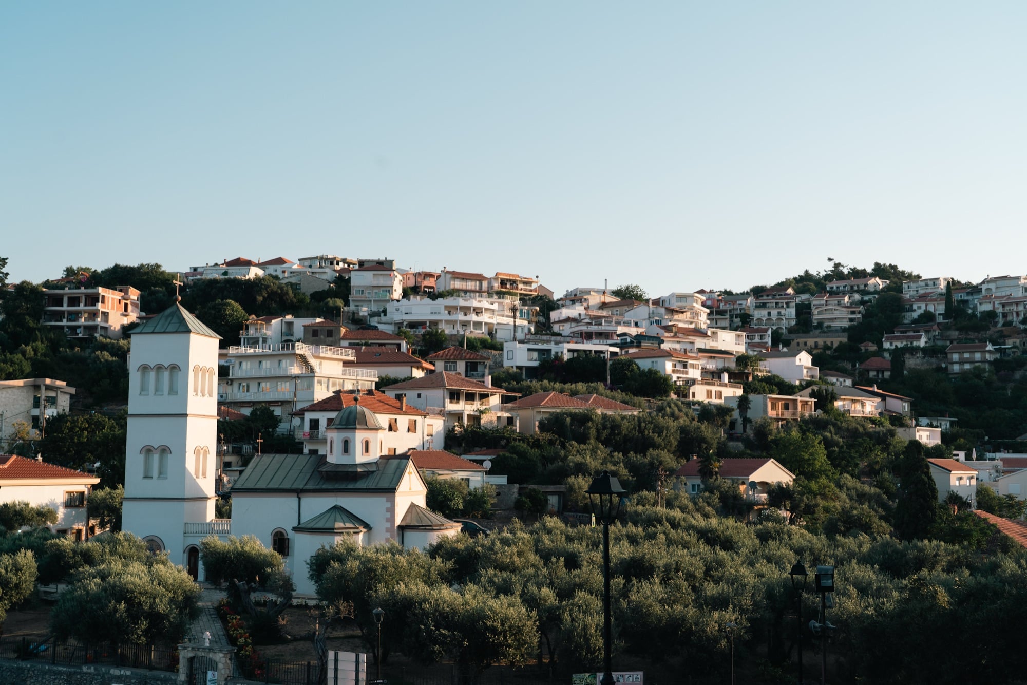 A white Orthodox church with a green-roofed bell tower amid olive groves below Ulcinj's houses stacked up the golden-lit hillside