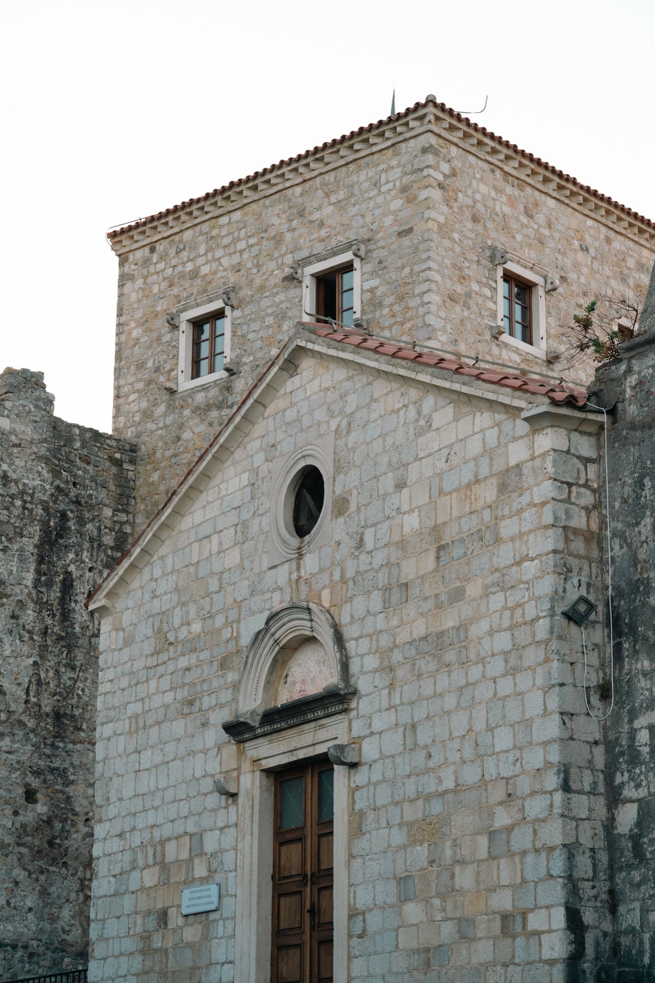 The weathered stone façade of Ulcinj's old-town church-turned-museum, a round window above its arched doorway beneath a tiled tower