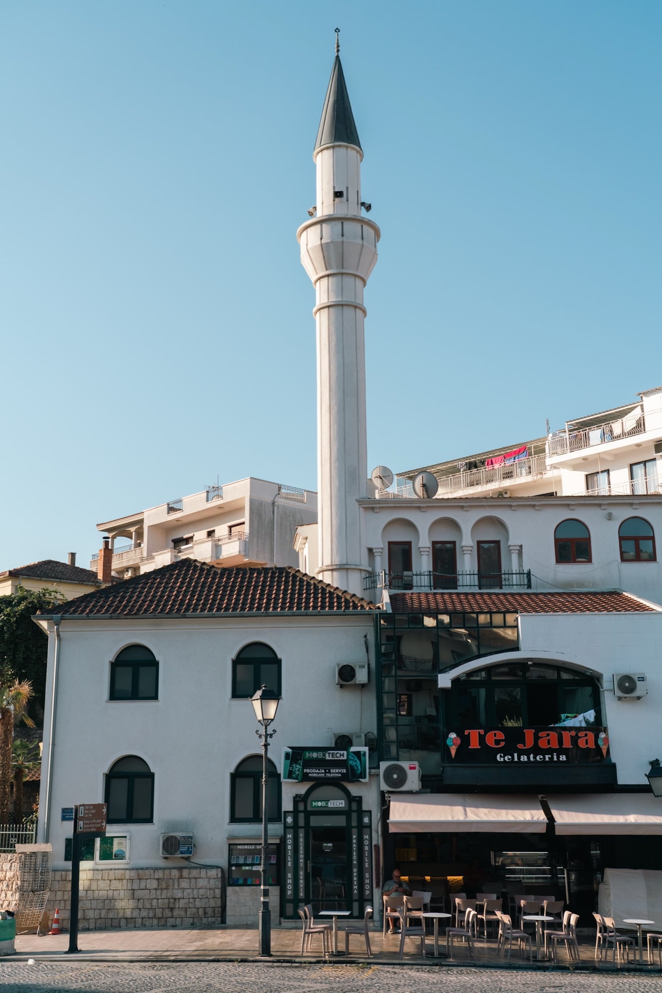 The white minaret of an Ulcinj mosque towering over the town square, a gelateria and cafés at street level below