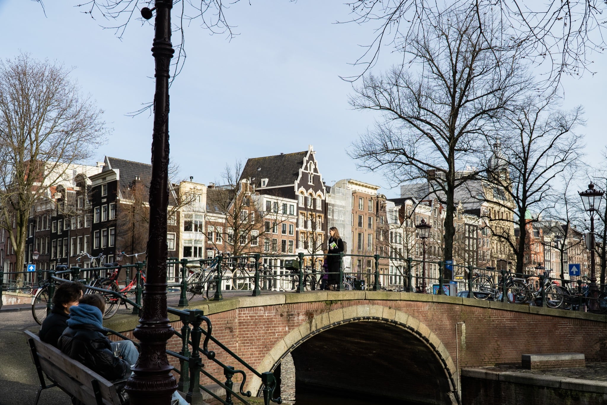 An arched brick bridge over a canal, a couple on a bench and gabled houses beyond